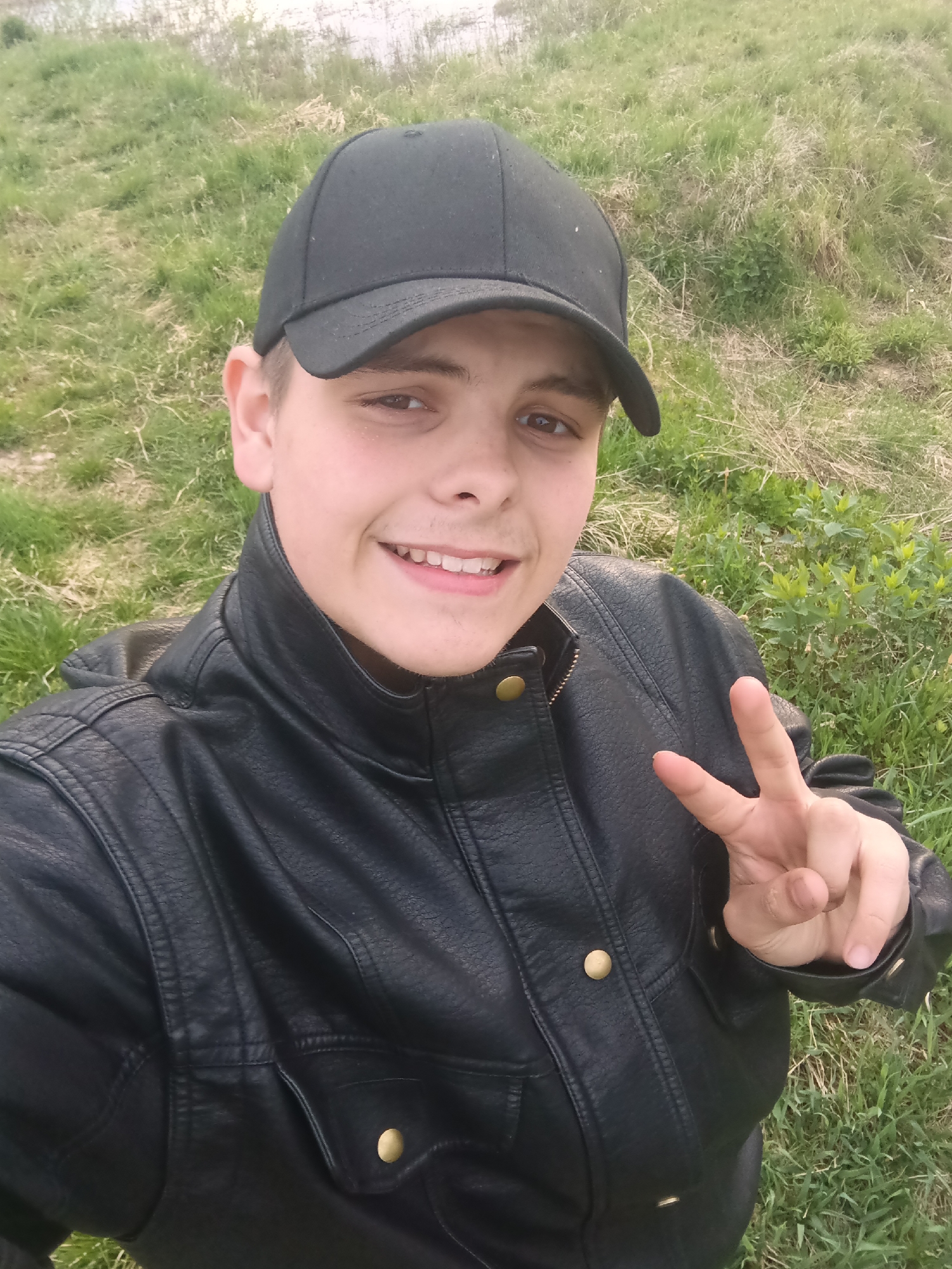 The image is a selfie of a young person wearing a black baseball cap and a black leather jacket. They are smiling and holding up a hand in a peace sign. The background is a grassy area with some vegetation. The image was most likely taken outside, and the presence of grass suggests a park or a field. The city of Красный Маяк, Russia, cannot be confirmed or denied based solely on the image.