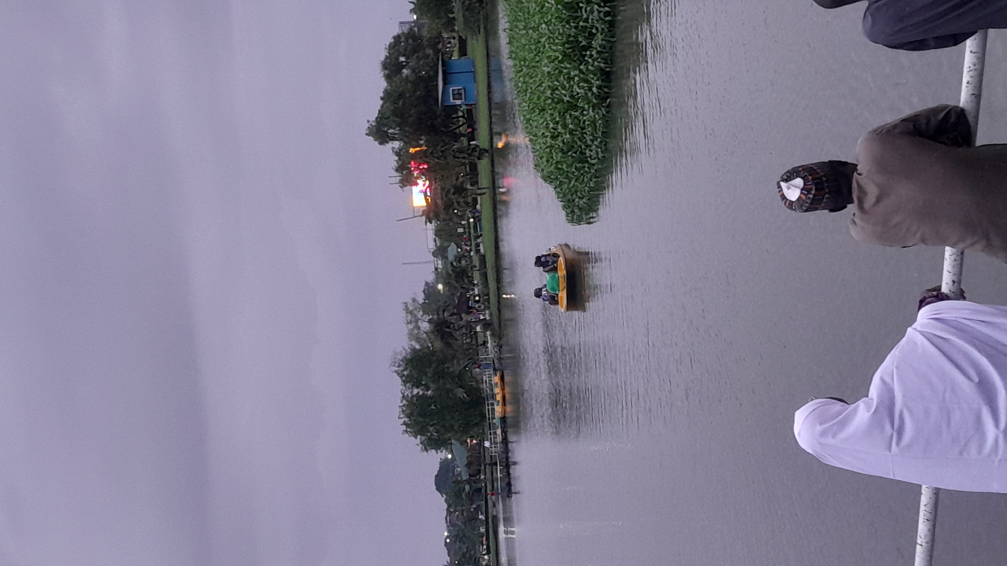 The image depicts a serene lake scene in the city of Rumuomasi, Nigeria. In the foreground, two individuals, one wearing a white hijab and the other a patterned cap, lean on a white railing, gazing out at the water. A yellow pedal boat with two occupants is visible in the middle of the lake. The far side of the lake is lined with lush green trees and a few structures, including a small blue building. The sky is overcast, with a hint of distant lights from a brighter area, suggesting it might be late afternoon or early evening. The overall atmosphere is calm and relaxed.