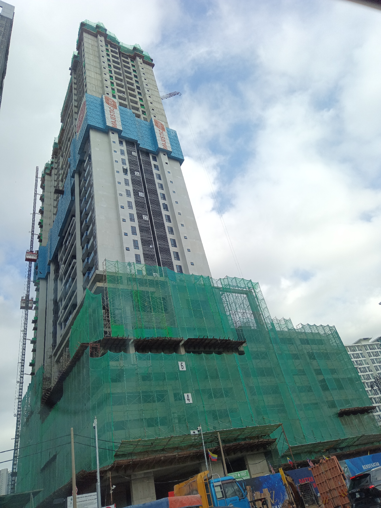 A low-angle, eye-level shot captures a colossal skyscraper under construction in Kuala Lumpur, Malaysia, against a backdrop of a partly cloudy sky. The immense concrete structure, shrouded in emerald green netting, dominates the frame. The netting, a ubiquitous safety feature at construction sites, conceals much of the building's lower and mid-levels, giving it a colossal, unfinished appearance. Blue banners with the text "MAJESTICA GEN" are visible on the upper sections of the building, indicating the project's name or developer. Cranes, vital to the construction process, are positioned on the sides of the skyscraper.
The building's facade features rows of dark-tinted windows, some of which are visible through gaps in the netting. The scale of the skyscraper is emphasized by the inclusion of external construction elevators, resembling vertical tracks with attached cages, that ascend the building's exterior. The lower portion of the image reveals a busy street scene. Various construction vehicles, including a blue truck and what appears to be a yellow excavator, are present. Numerous traffic barriers and signage are also visible, suggesting an active urban environment surrounding the construction site. The presence of Malaysian flags and other signage indicates the project is located within Malaysia. The overall impression is one of dynamic urban development, highlighting the rapid growth and ongoing construction projects characteristic of Kuala Lumpur.