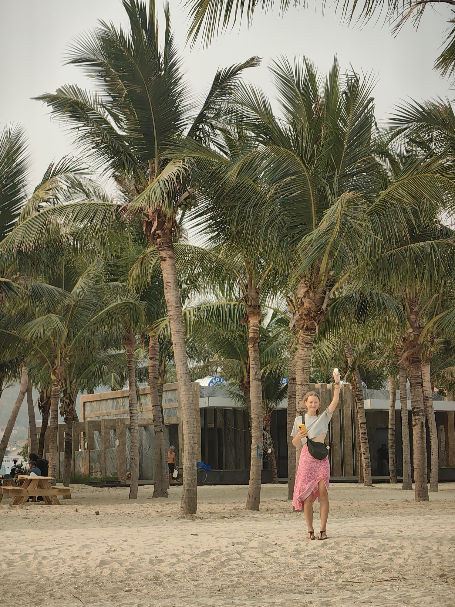 A woman stands on a sandy beach in Bai Chay Ward, Vietnam. She is fair-skinned, wearing a gray crop top, a pink floral skirt, and black sandals. She holds a phone in one hand and raises the other. Behind her are palm trees and a structure resembling a building under construction. In the background, people are seated at a picnic table, and another individual is walking nearby. The sky is overcast, and the sand is light-colored.