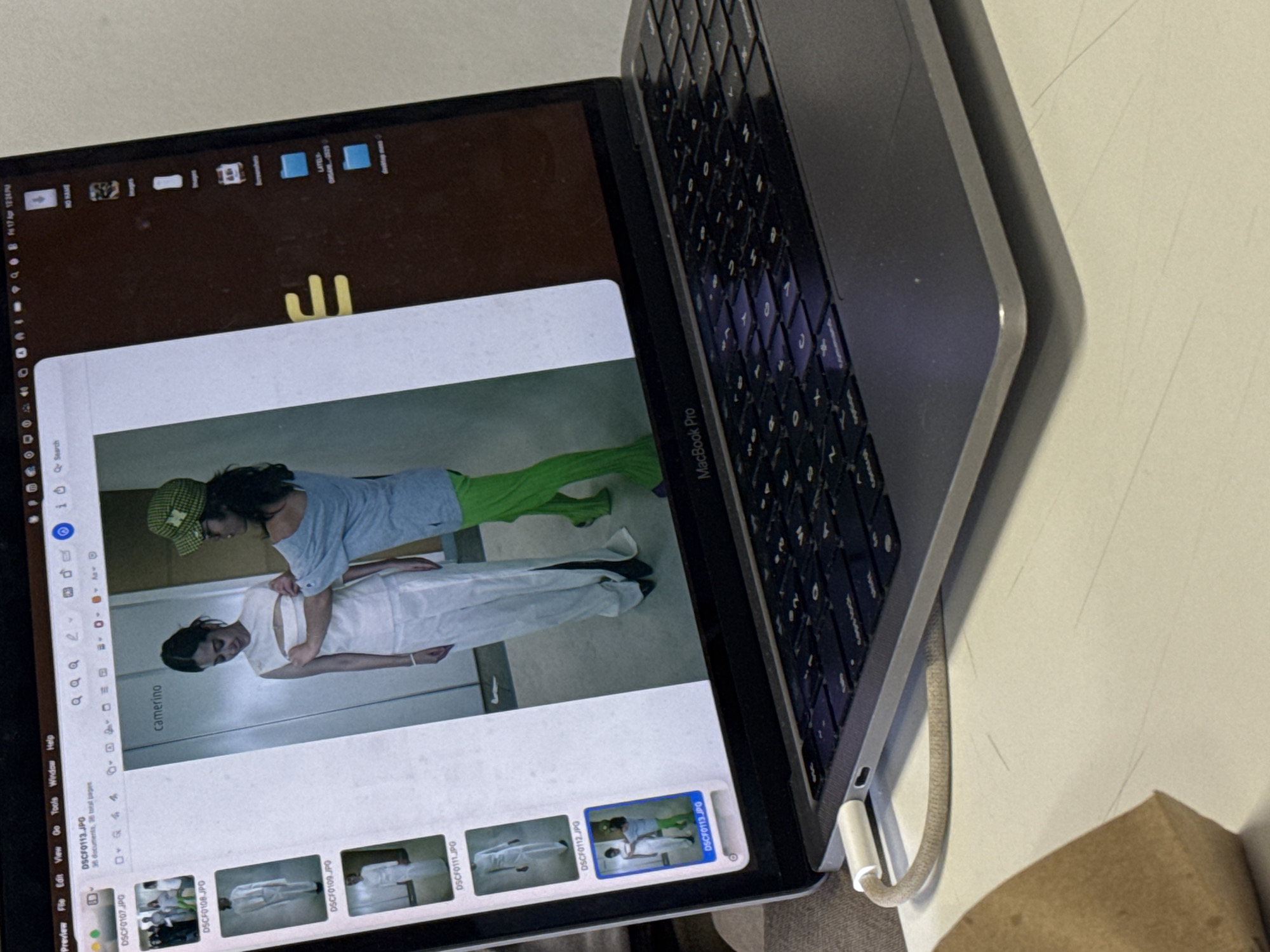Here is a detailed description of the image:
The image shows a MacBook Pro laptop displaying an image in Preview. The image appears to depict two people in a clinical or studio environment. One person, wearing a green and yellow hat and a blue shirt and green pants, appears to be adjusting the white garment of the other person.
The laptop screen shows icons and a search bar. The dock at the bottom displays several thumbnails of image files.
The laptop is placed on a white table, and a charging cable is plugged into the side. A cardboard object is to the right. The image is taken at an angle, providing a partial view of the keyboard.