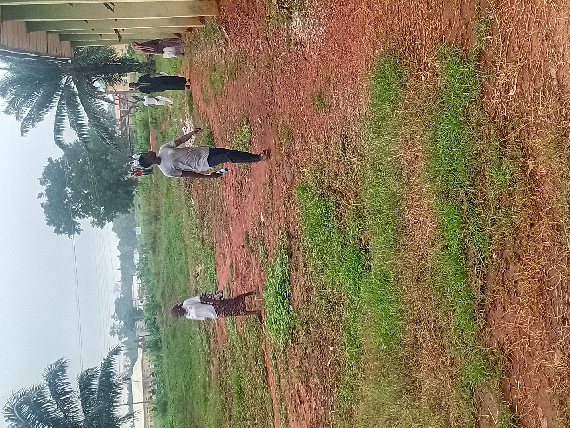A group of people are walking on a dirt path in front of a building with a pale green exterior. The path is lined with patchy green grass and brown earth. In the foreground, a young man in a grey t-shirt and dark pants walks towards the right. Further down the path, a woman in a white t-shirt and a patterned skirt walks towards the left. Several other people, some in traditional attire, are visible in the background walking on the path. Tall palm trees are present on the left side of the image. The sky is overcast.