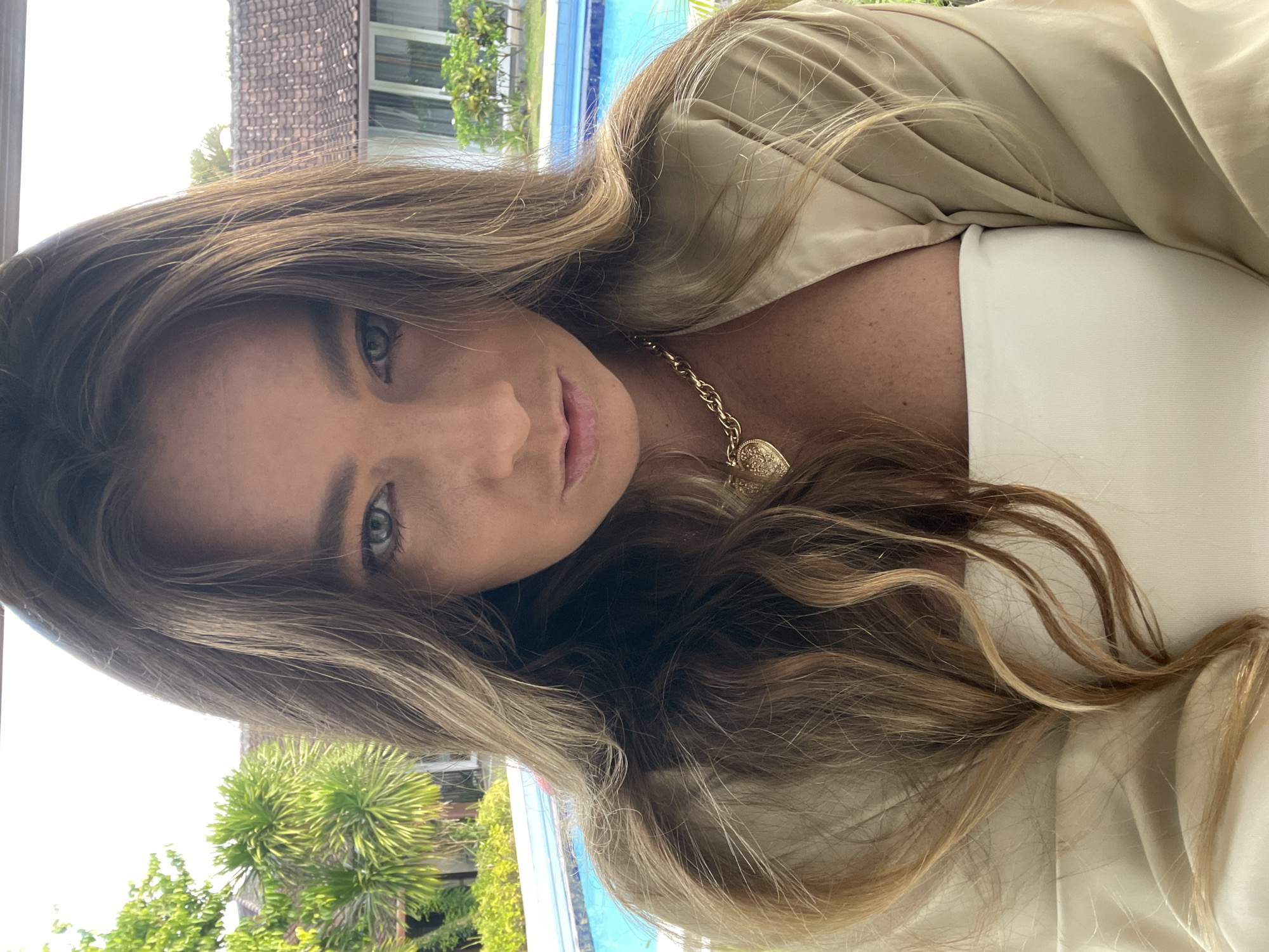 A medium shot, front-facing photograph shows a woman with long, wavy brown hair and blue eyes. She is wearing a white strapless top and a tan open-fronted shirt. A gold necklace with a heart-shaped pendant is visible around her neck. She is looking directly at the camera with a serious expression. The background features a swimming pool and lush green foliage, characteristic of a tropical resort setting in North Kuta, Indonesia. The lighting suggests it is daytime, with natural light illuminating the scene.
