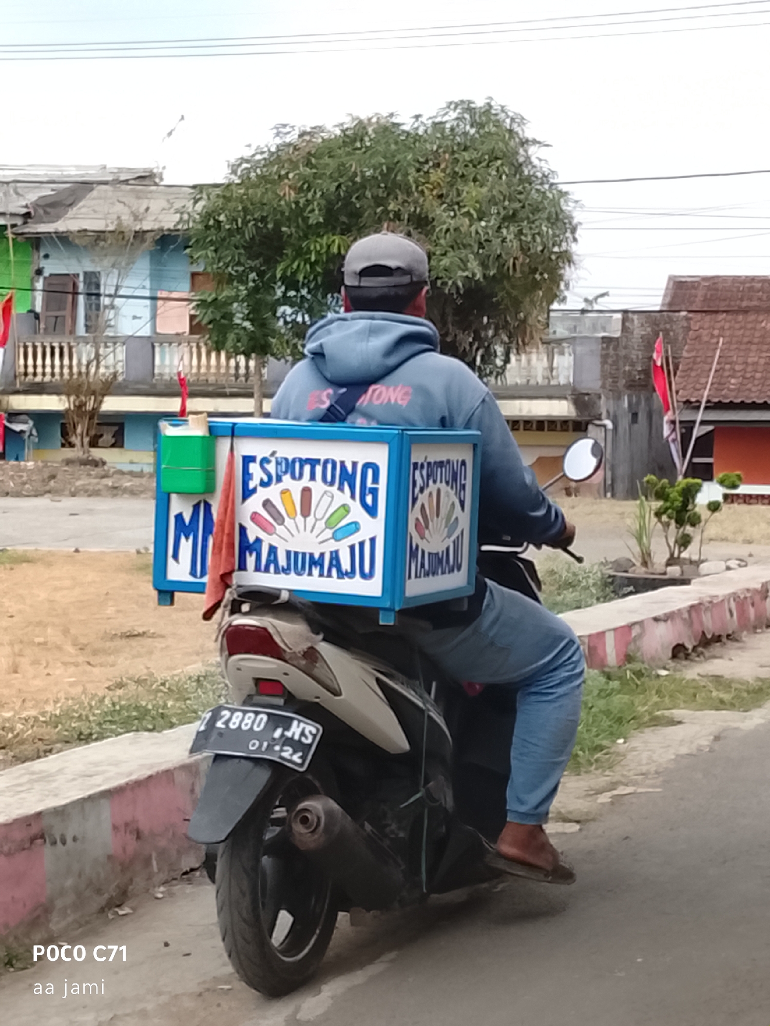 The image shows a man riding a motorcycle with a large box on the back, likely used for selling ice cream. The box is blue and white and has the text "ES POTONG MAJUMAJU" written on it, along with an illustration of several ice pops. The man is wearing a blue hoodie and blue pants, and a grey cap. He is wearing a sandal on his right foot. The motorcycle has a license plate that reads "2880 NS". In the background, there are several buildings, trees, and a sidewalk. The overall scene appears to be in a residential or commercial area in Tasikmalaya, Indonesia, based on the writing on the box and the style of the buildings.