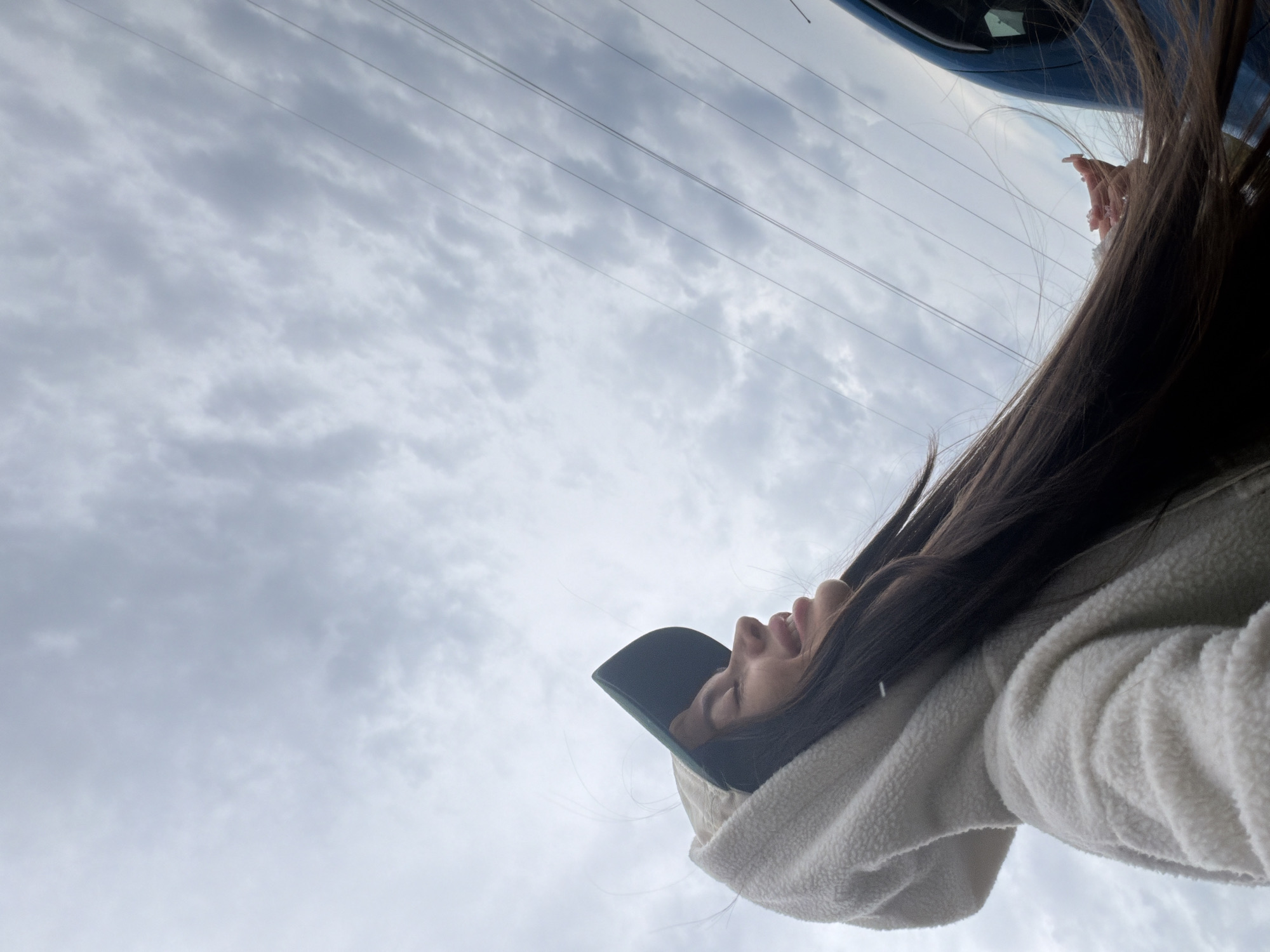 A young woman is captured in a low-angle shot, her head tilted back and eyes closed as she smiles upwards at a cloudy sky. She is wearing a cream-colored fleece hoodie with the hood up and a baseball cap with a green brim and a lighter colored crown. Her long, dark hair is blowing around her face and shoulders. In the background, thin wires stretch across the sky, and a portion of a dark blue vehicle is visible on the right side of the frame. The overall lighting suggests an overcast day. The setting is outdoors, with the sky and electrical wires being prominent environmental features.