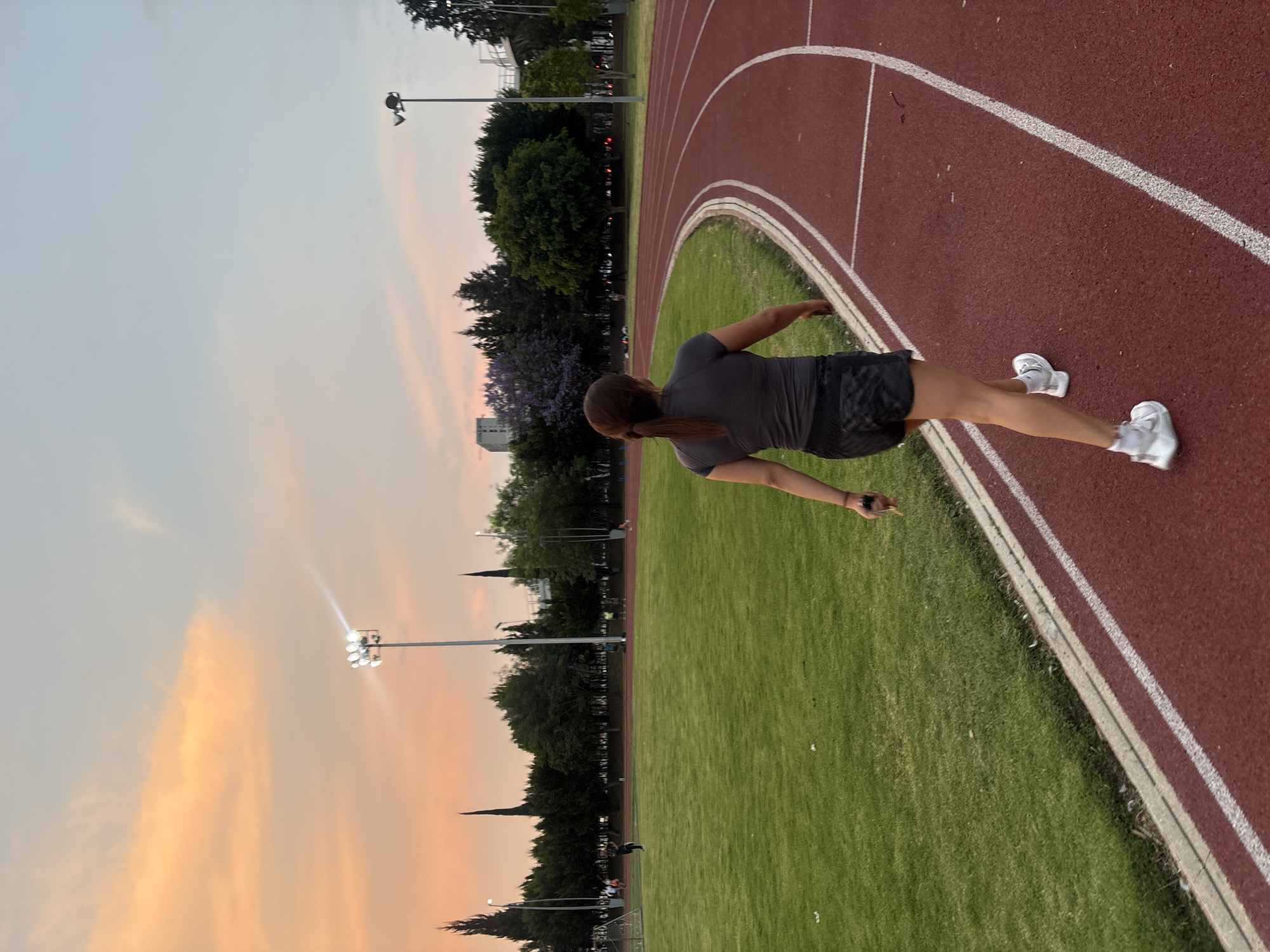 An outdoor athletic track scene is captured at dusk or sunset in Puebla, Mexico. The image is oriented with the horizon on the left, but describes a standard landscape view. A single person, seen from behind, is walking or jogging on the red running track. The individual has dark hair tied in a ponytail and is wearing a dark grey short-sleeved top, dark patterned athletic shorts, and white athletic shoes. Their arms are slightly bent, suggesting active movement.
The track itself is red with distinct white lane markings. Inside the track's curve lies a vibrant green grass field. In the background, tall stadium light poles are visible, with some lights illuminated, indicating the diminishing daylight. A line of dense trees, some displaying purple blossoms (possibly jacaranda trees), borders the track. Distant buildings and structures are faintly visible against the horizon. The sky is a prominent feature, displaying warm gradients of orange, pink, and soft blue, characteristic of a sunset. The setting conveys a serene atmosphere for evening exercise. There is no visible text in the image.