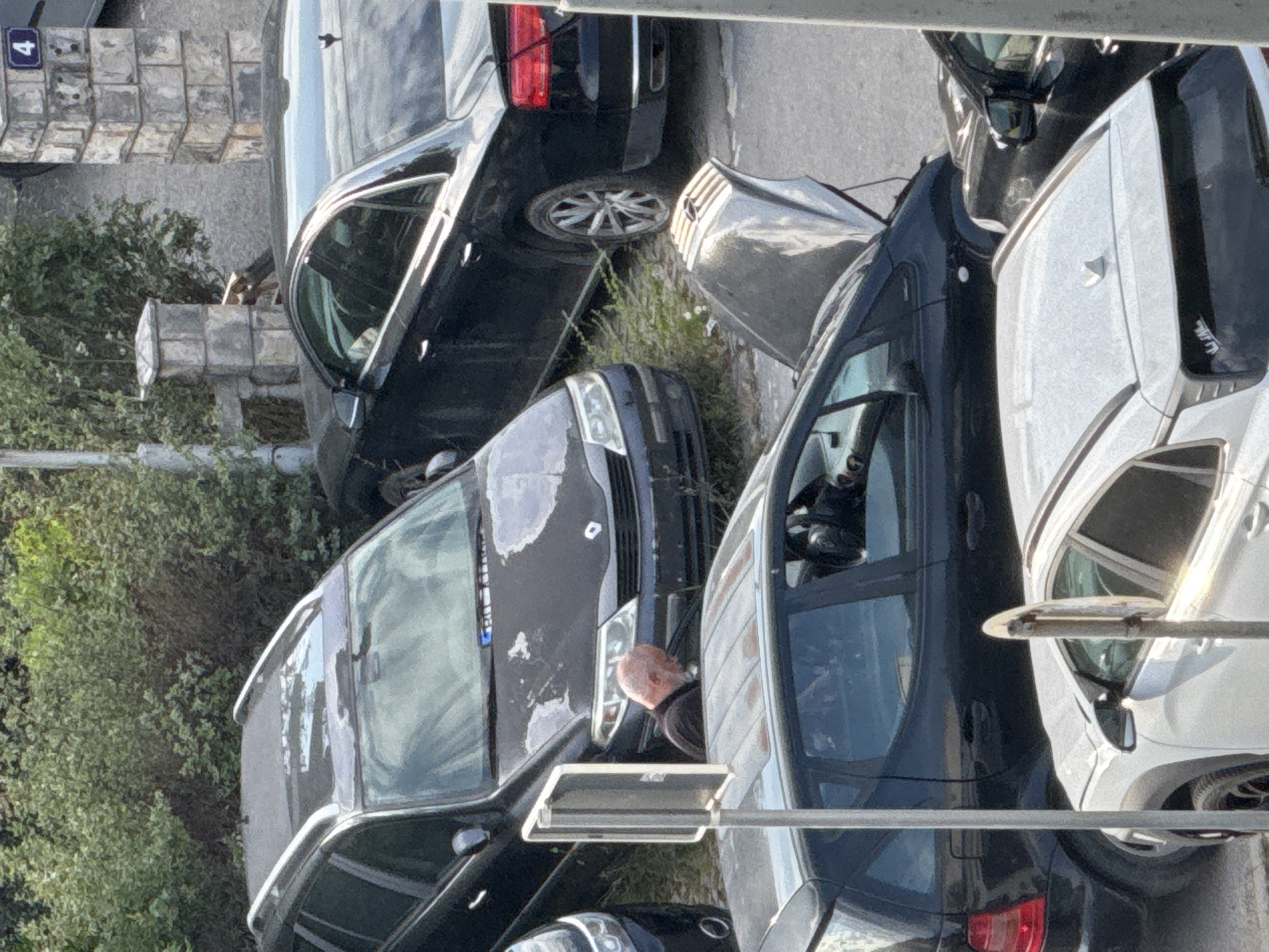 An overhead, medium-shot captures a street scene in Budva, Montenegro, with multiple parked cars and a man attending to one of them. The foreground is dominated by the roof and side of a white SUV. To the left, a black station wagon is parked, its paint visibly peeling on the roof. A man with a bald head and a beard, wearing a dark shirt, is positioned between the station wagon and another black car, with his upper body obscured by the station wagon's roof. He appears to be looking downwards, possibly working on the vehicle. The station wagon has its hood up. Further back, a black sedan is parked, and to its right, another black sedan with its hood open is visible. The environment suggests an urban setting, with a stone pillar and overgrown greenery. The license plate of the station wagon is partially visible. A blue sign with the number "4" is affixed to a stone pillar in the background.