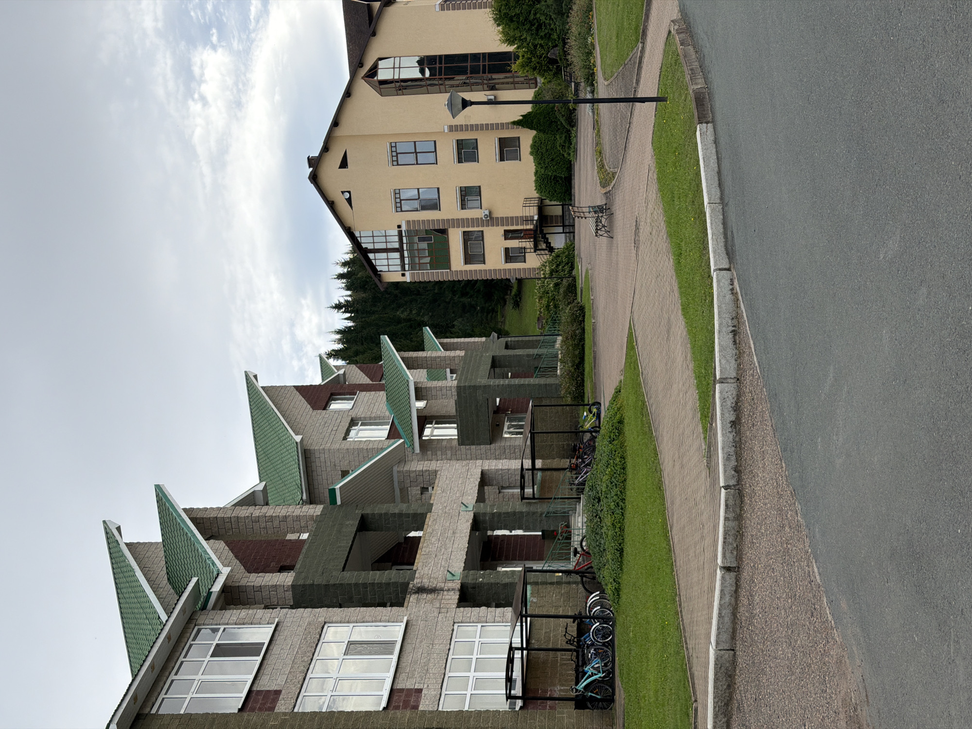 The image depicts a residential complex with modern architecture. In the foreground, a paved road curves to the right, bordered by a gravel area and then a concrete curb. Beyond the curb is a patch of green grass. To the left, a row of buildings with light-colored brick facades and green, angular roofs are visible. These buildings have multiple floors with large windows. In front of these buildings, there are covered bike racks with several bicycles parked beneath them. A small, overgrown green bush is located in front of one of the bike racks.
To the right of the brick buildings, a taller, cream-colored building with a dark brown roof stands out. This building also features multiple windows, some of which are large and appear to be a reflective glass. A black lamppost is situated in the paved area in front of this building. In the background, a dense forest of evergreen trees covers a hillside. The sky is overcast with scattered clouds. There are no people visible in the image. The location is identified as the city of Assy, Russia.