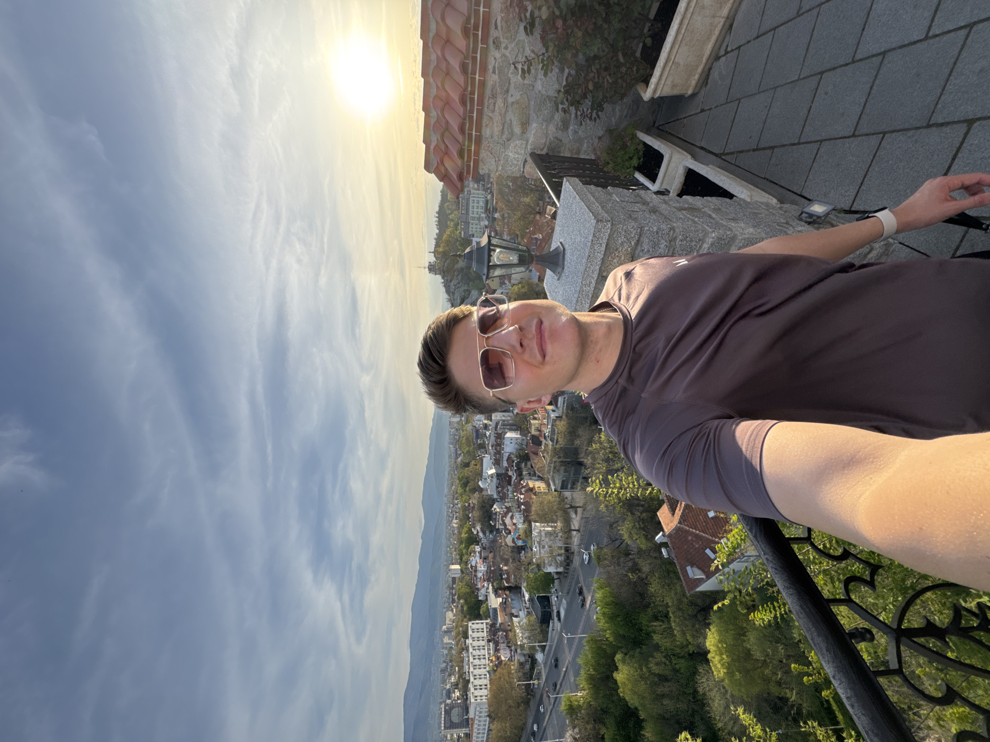 A man wearing sunglasses and a brown t-shirt is taking a selfie. He is standing on a balcony overlooking the city of Plovdiv, Bulgaria. The buildings in the city appear to be a mix of residential and commercial buildings, and there are many trees and green spaces. In the background, there are hills. The sky is bright and sunny, with some clouds. The balcony has a stone base and a black metal railing. A streetlamp is visible on the balcony. The man is looking at the camera and smiling. The man is likely at the top of Nebet Tepe in the Old Town of Plovdiv.