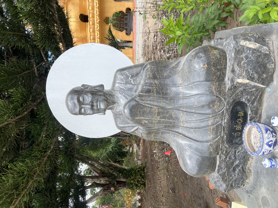 An outdoor scene in Đăk Mar, Vietnam, features a dark grey marble statue of a seated, bald male figure, likely a Buddhist patriarch, positioned centrally. The statue depicts an elderly individual with serene facial features and hands clasped, wearing flowing robes; the stone exhibits gold-colored veining. A large, flat white circular disc serves as a halo behind the figure's head. The statue rests on a multi-tiered dark grey stone base, with "TỔ ĐỆ-CA-ĐA" inscribed in gold lettering on the front. At the base, a blue and white ceramic incense burner filled with ash is flanked by two smaller matching ceramic adornments. The immediate foreground consists of disturbed earth with sparse green vegetation, including small leafy plants and a shrub with red ribbons or blossoms. Red construction bricks are visible near the statue's base on the left. In the background, large trees with dense, dark green needle-like foliage are visible on the left. To the right, a prominent golden-yellow pagoda-style building with intricate arched openings and multiple tiers is situated on a cleared area.