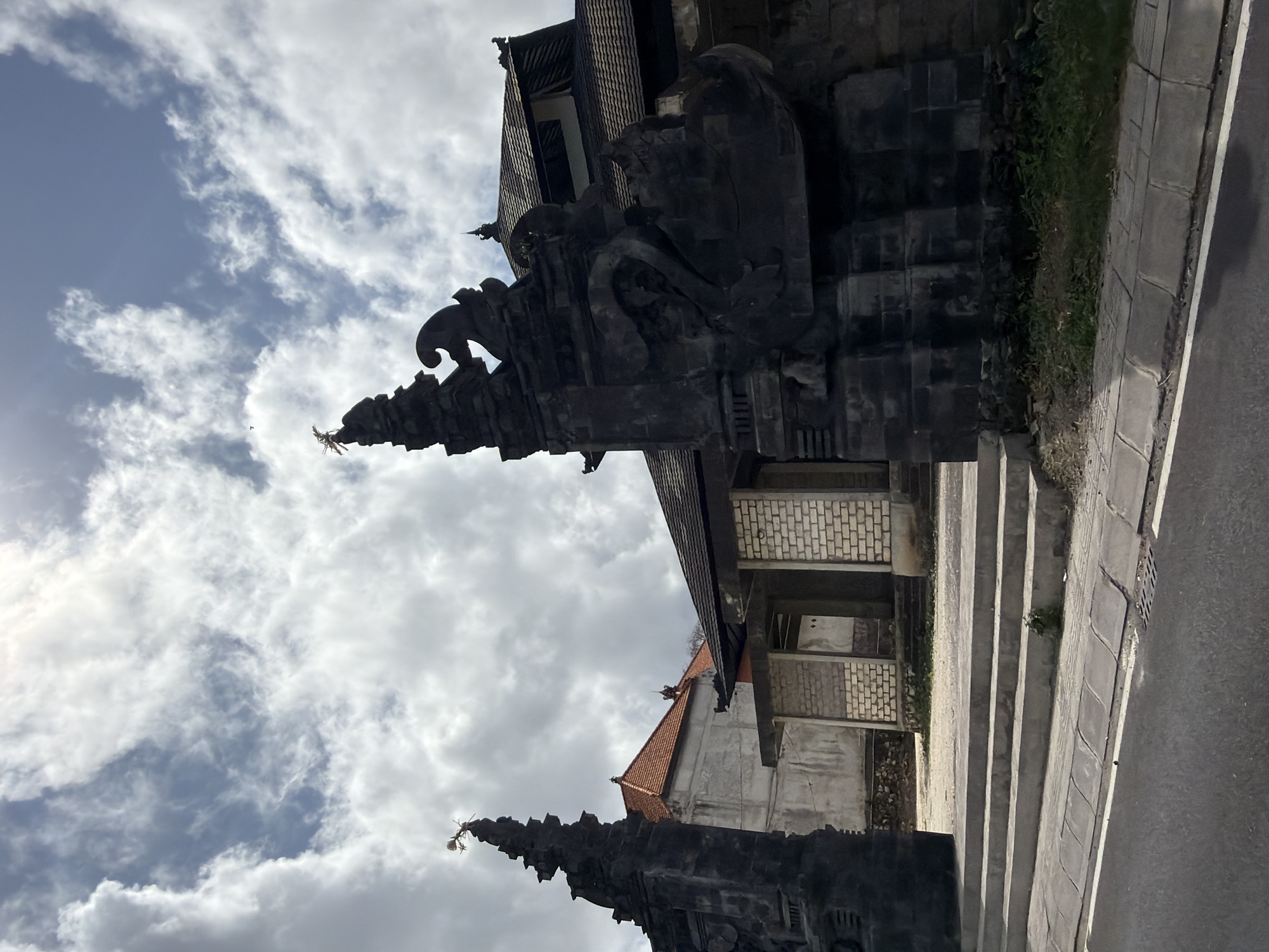 In Kuta Selatan, Indonesia, a traditional Balinese temple gate stands imposingly against a cloudy sky. The dark, stone structure is intricately carved with mythological figures and ornate detailing, featuring tall, pointed spires topped with metallic decorations. To the left of the main gate, another similar spire is visible. In the background, a building with a terracotta-tiled roof and concrete walls is partially obscured by the temple structures. A paved pathway with concrete steps leads up to the entrance of the temple, with a road in the foreground. The scene is captured from a low angle, emphasizing the grandeur of the temple gate.