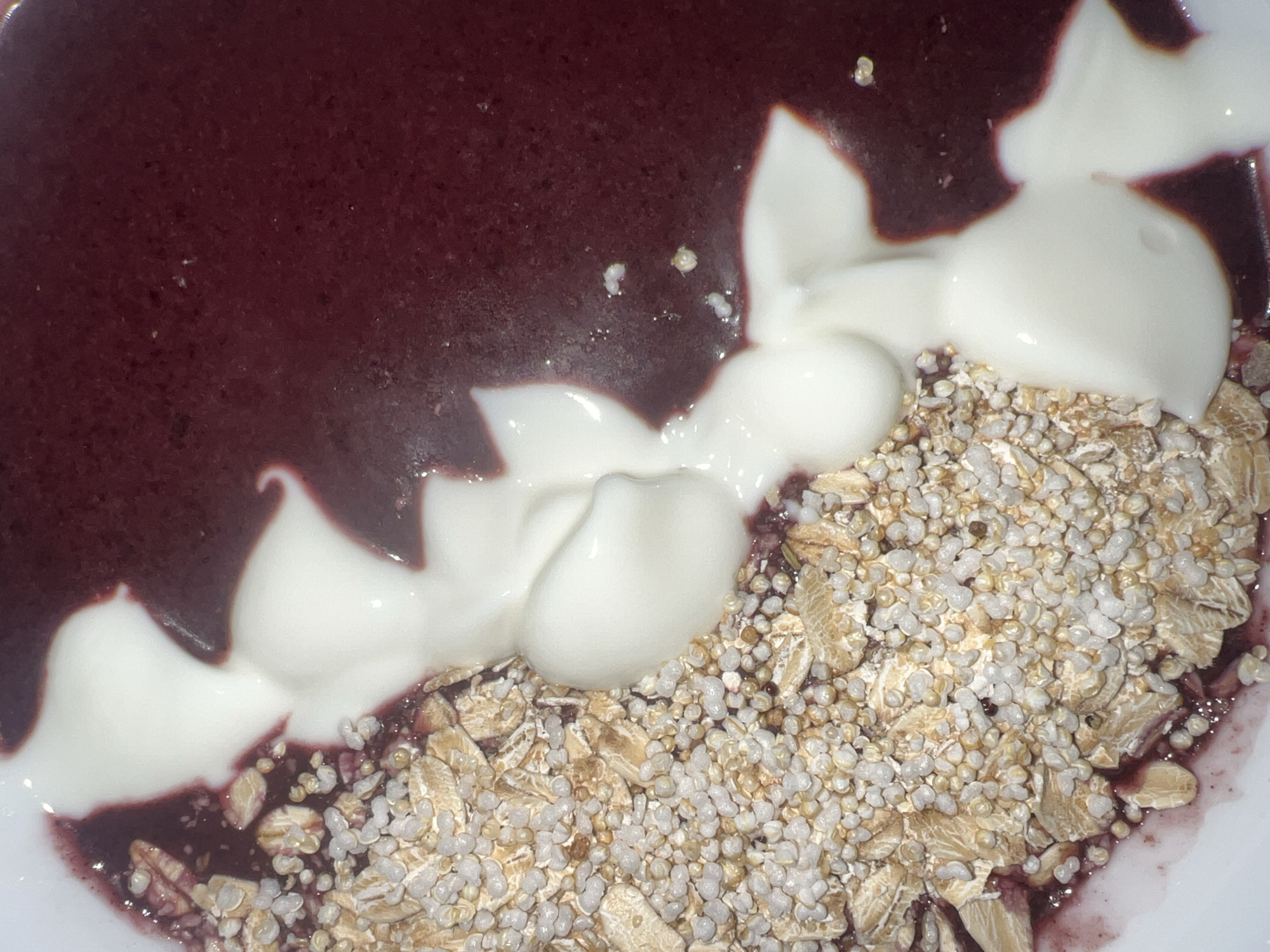 The image shows a bowl of food, likely a breakfast or snack bowl. The bowl contains a dark purple smoothie or puree on one side, with white yogurt swirls layered on top. Adjacent to the smoothie is a mixture of granola and puffed rice. The bowl is likely photographed in a domestic kitchen setting. The location cannot be determined from the image.