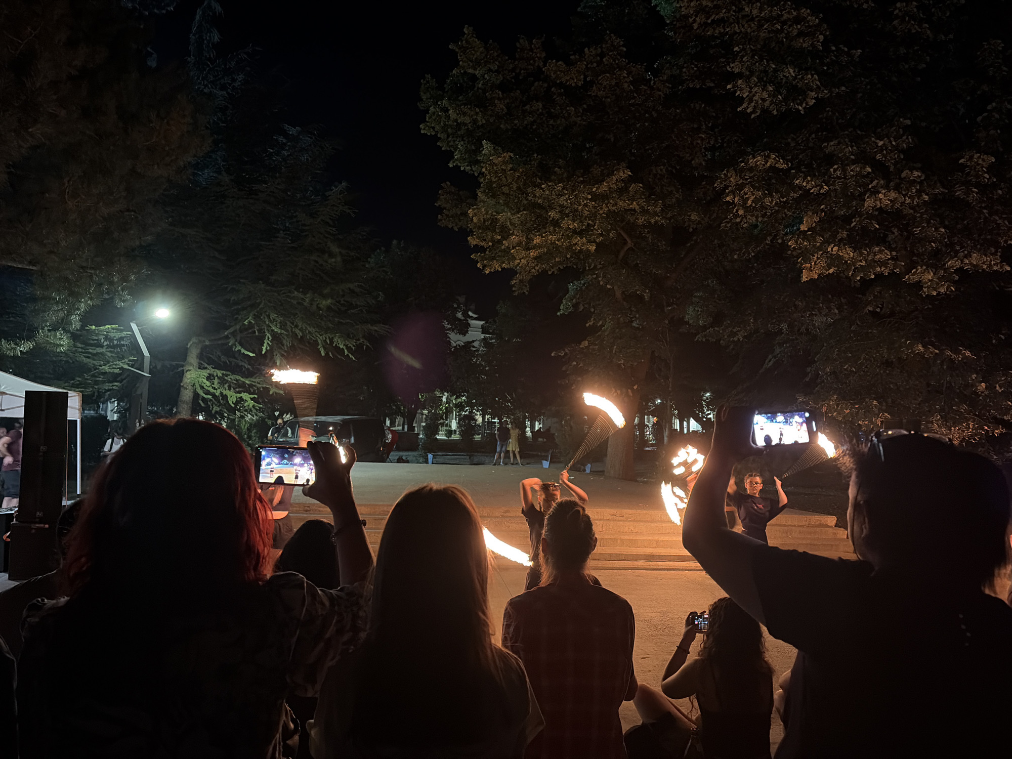 This image captures a night-time outdoor fire performance in the city of Vratsa, Bulgaria. In the foreground, several spectators are silhouetted against the bright flames, with some holding up smartphones to record the event. The performers, visible in the mid-ground, are actively engaged in manipulating fire, creating dazzling visual displays with flaming props. The background is dominated by trees and streetlights, indicating a park or public square setting. The overall atmosphere is one of energetic entertainment and audience engagement.
