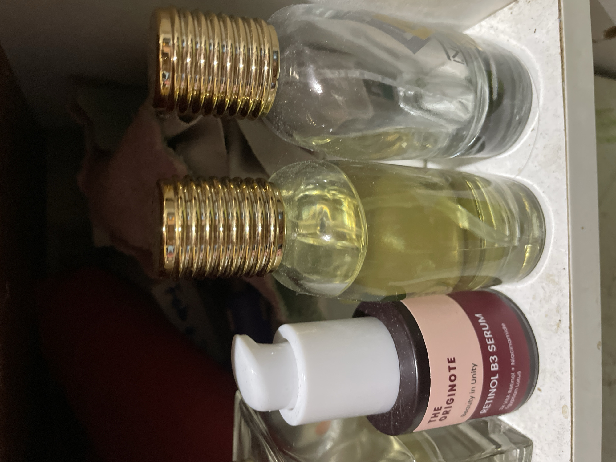 The image displays a close-up, top-down view of several beauty or personal care products arranged horizontally, likely inside a drawer or cabinet in Davao City, Philippines.
In the foreground, there are three bottles. Two of these are clear glass bottles with distinctive ribbed, gold-colored caps. The uppermost of these two contains a clear liquid, while the one below it holds a yellowish liquid. Both appear to be fragrances or similar liquid products.
Below these two bottles is a third, smaller, dark-colored bottle fitted with a white pump dispenser. This bottle has a label wrapped around it. Clearly visible on the label, from top to bottom, are the words "THE ORIGINOTE" followed by "Beauty in Unity." Below this, "RETINOL B3 SERUM" is prominent, with smaller text underneath that reads "2% AHA Retinol + Niacinamide" and "Egyptian Lotus."
The bottles are resting on a light-colored, possibly white or off-white surface that shows some dust or debris. To the left side of the frame, a dark reddish-brown fabric or object is partially visible in the blurred background. The overall lighting suggests an indoor setting, possibly with artificial light, without any specific time of day indicated. No activity is depicted in this still life arrangement of products.