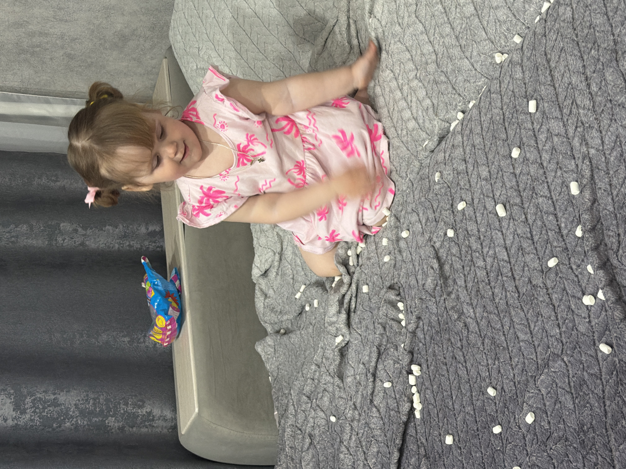 A young child with fair skin and blonde hair styled in pigtails stands on a light gray upholstered sofa in what appears to be an indoor setting. She is wearing a light pink dress with a pink palm tree pattern. The sofa is covered with a gray textured throw blanket with small white objects scattered across it. A colorful bag is visible on the sofa cushion near a gray curtain or wall. The location is Chaikovsky, Russia.