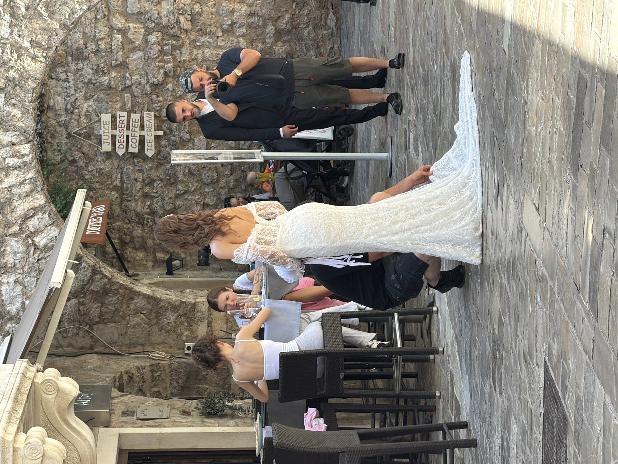 The image depicts an outdoor scene in the city of Budva, Montenegro, possibly near a restaurant or cafe. In the foreground, a woman in a white wedding dress is being photographed. A photographer, wearing a black t-shirt and a camouflage cap, is actively taking pictures of her. To the left, two women are seated at a high table, one facing away from the camera and the other looking down. Behind them, a person is partially visible with a baby in a stroller. The background features a stone wall with signage indicating "COFFEE TOO" and a directional sign with options like "JUICE," "DESSERT," "COFFEE," and "ICE CREAM." The ground is paved with cobblestones. The overall atmosphere suggests a candid moment captured during a wedding photoshoot or a casual gathering.