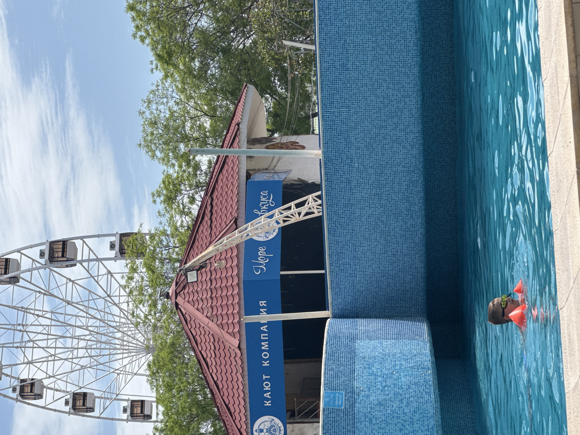 The image depicts a swimming pool with a child wearing goggles and arm floaties in the water. Adjacent to the pool is a structure with a red-tiled roof and a blue sign that reads "КАЮТ КОМПАНИЯ Море Вкуса" in Cyrillic text. In the background, a Ferris wheel is visible against a sky with scattered clouds. This scene is located in the city of Yevpatoriya, Ukraine.