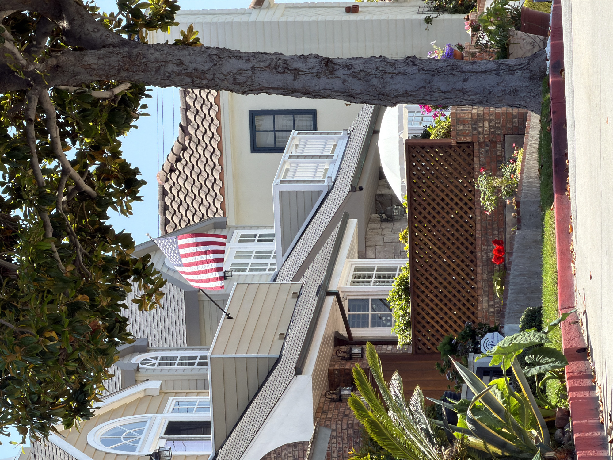 The image shows a residential street in Newport Beach, California. The houses are multi-story, with varying architectural styles and materials, including wood siding, brick, and tile roofs. One house has an American flag displayed. A mature tree is partially visible, obscuring some of the houses. There is a sidewalk and a landscaped area with plants and flowers bordering the street.