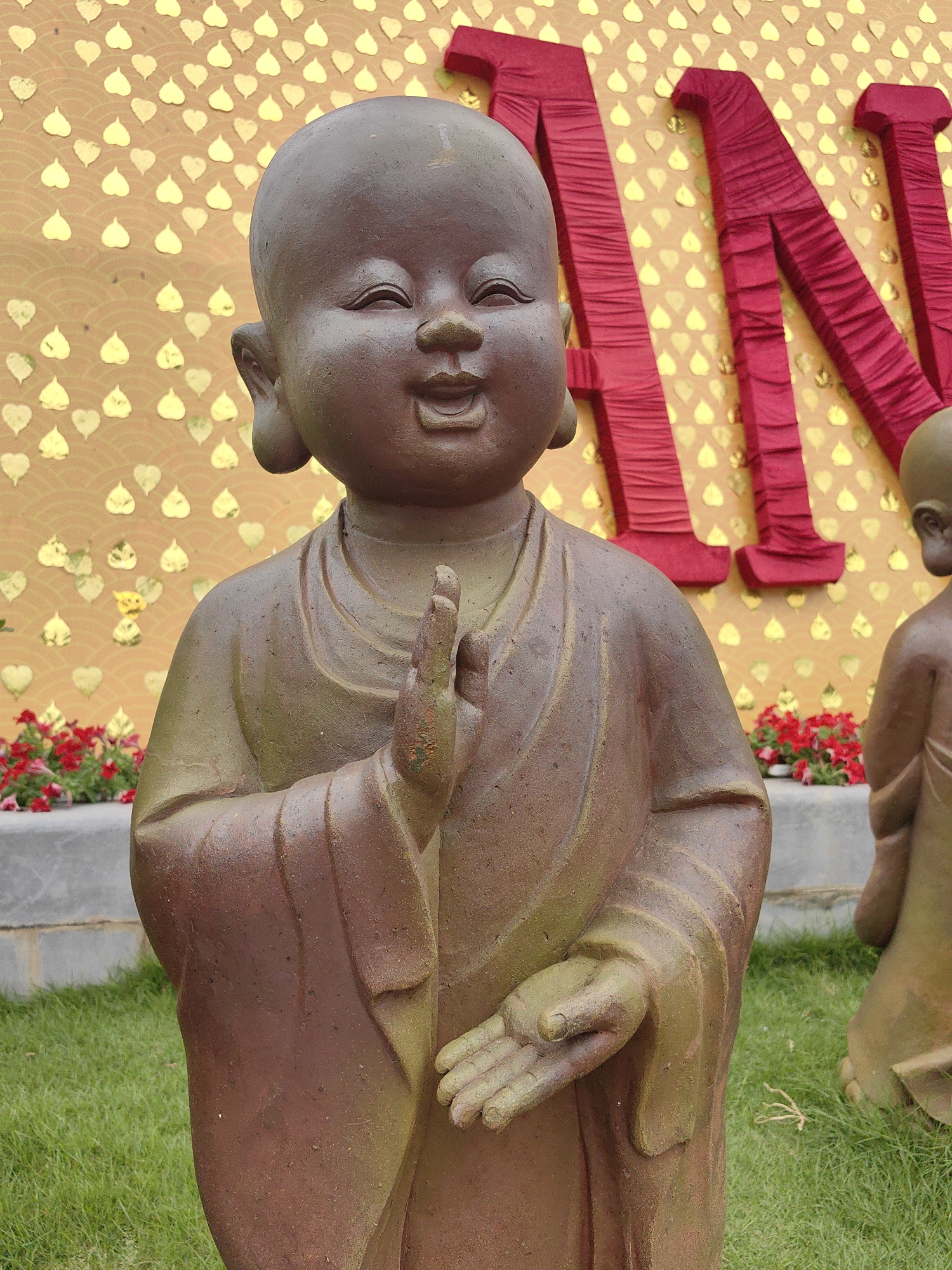 A serene outdoor scene is captured, focusing on a statue of a young Buddhist monk (a novice or "little monk") made of a reddish-brown material with subtle green highlights. The monk's face is round, with closed eyes suggesting a peaceful smile, and large, elongated earlobes. One hand is raised in a teaching gesture (possibly vitarka mudra), while the other is open, palm facing upwards, in a gesture of giving or receiving. The statue wears simple robes that drape naturally.
To the right, partially visible, is another similar statue, possibly seated or kneeling, reinforcing the religious theme.
The background features a vibrant golden-yellow wall adorned with numerous small, golden, heart-shaped or Bodhi leaf-shaped motifs, arranged in a subtle wavy pattern. Against this wall, large, three-dimensional letters, wrapped in rich red fabric, spell out part of a word. The letters "AN" are clearly visible, with portions of a preceding letter (possibly 'M' or 'N') and a succeeding one.
In the midground, below the red letters and behind the main statue, are grey concrete planters filled with clusters of small, vivid red and pink flowers. The foreground and surrounding area consist of well-maintained, bright green grass. The scene is brightly lit, suggesting it is daytime. The overall impression is one of tranquility and cultural celebration, likely in a temple garden or a public space in Hong Gai Ward, Vietnam.