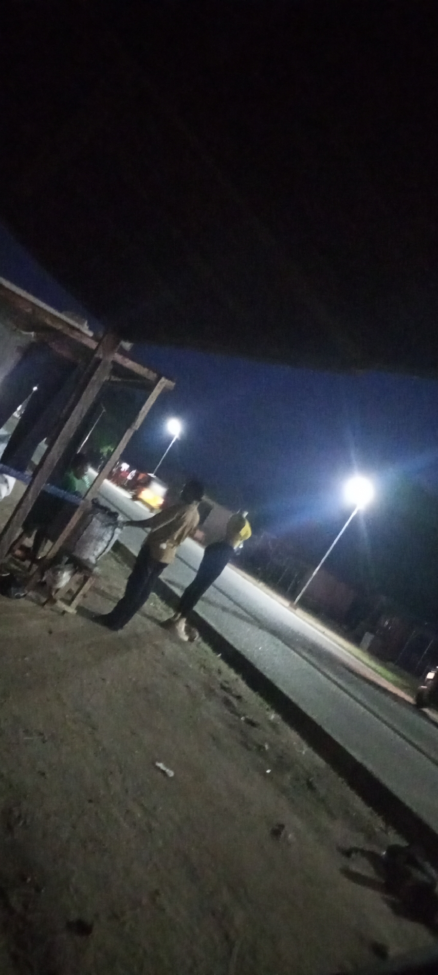 In Jalingo, Nigeria, at night, two individuals are standing near a roadside structure. One person, wearing a light-colored shirt and dark pants, is facing left, while the other, in a yellow shirt and dark pants, is facing right. The scene is illuminated by streetlights. The area appears to be a dirt road adjacent to a paved road, with a simple wooden structure to the left. Some debris is scattered on the ground. The sky is dark blue, indicating nighttime.