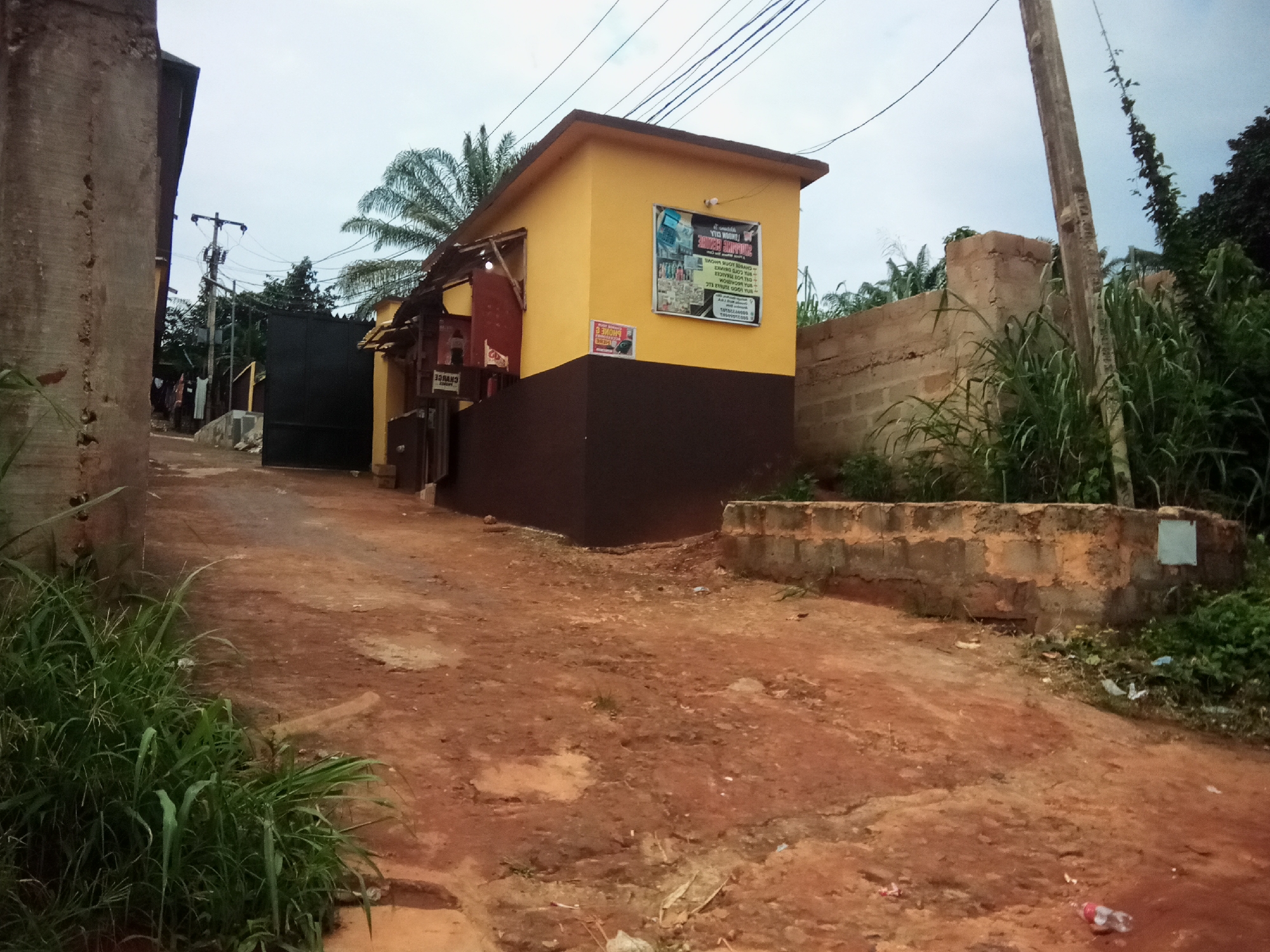 This image depicts a dusty, unpaved road in Oko Town, Nigeria, leading towards a small, yellow and brown building. The building appears to be a commercial establishment, possibly a shop or service center, as indicated by a sign on its facade advertising "SUMMIT CONNECT PHONE" and other services. A black metal gate stands open to the left of the building, suggesting it might be an entrance to a compound. The environment is characterized by overgrown vegetation, a tall utility pole, and a low brick wall. The sky is overcast, contributing to a diffused light. There are no people visible in the scene. The overall impression is one of a typical urban fringe setting in Nigeria.