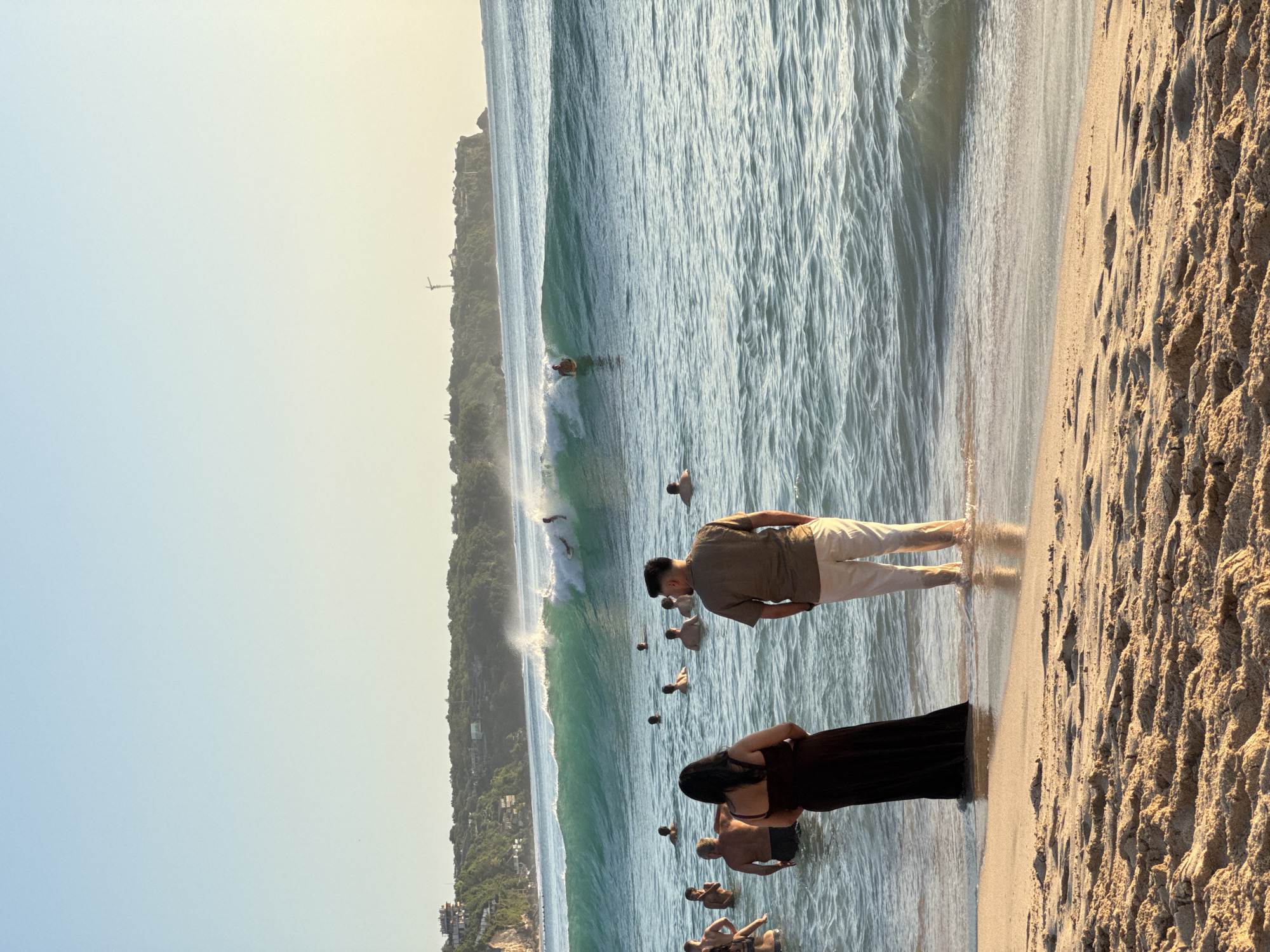 This image shows a beach scene in Kuta Selatan, Indonesia. The beach is crowded with people wading in the water, with others appearing to swim or surf further out. There is a large wave breaking in the background. Two people are standing on the sand, observing the activity; one is wearing a tan shirt and white pants, and the other is wearing a long black dress. The sand is textured with footprints. In the distance, a cliff covered with vegetation and buildings is visible against a clear sky.