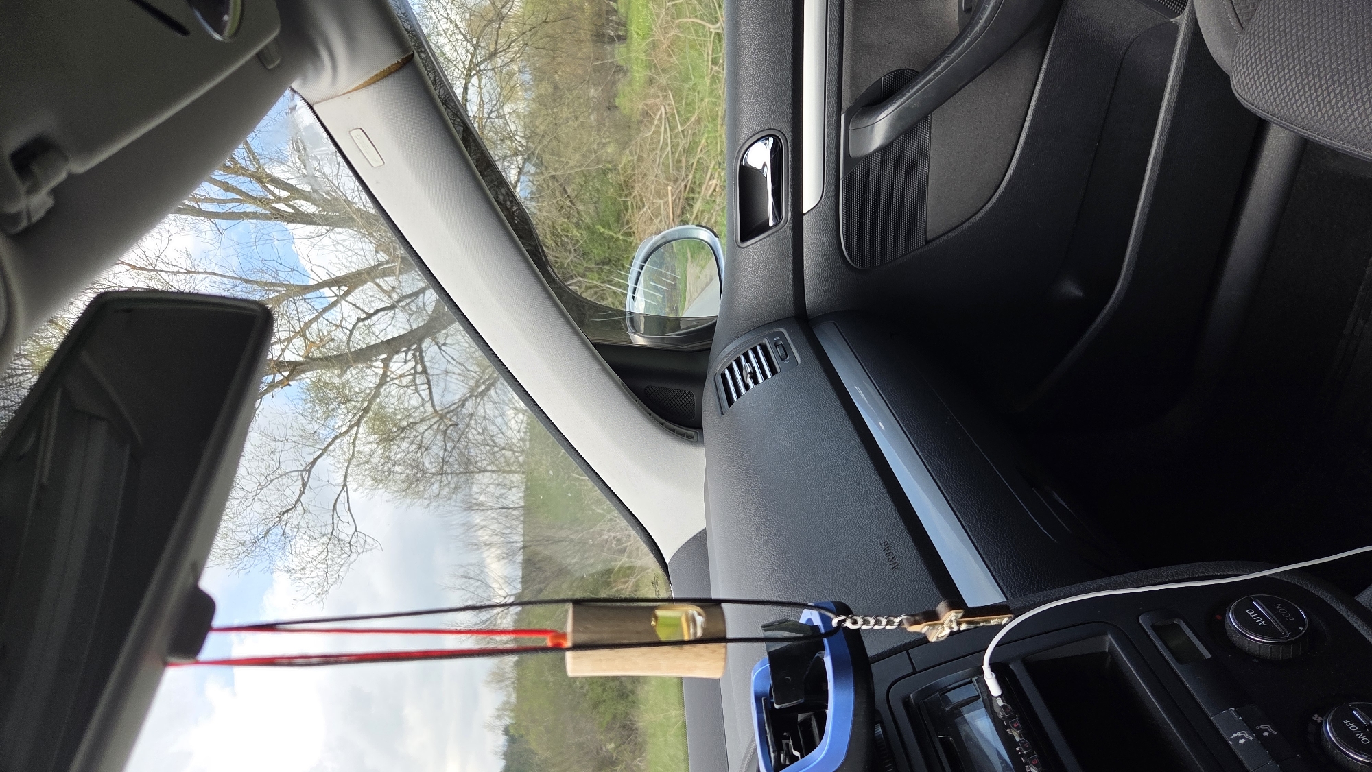 The image displays the interior of a car, viewed from the front passenger seat looking towards the windshield, dashboard, and passenger door. The original image is oriented vertically but should be viewed as a landscape photo rotated 90 degrees counter-clockwise.
Inside the car, the dark grey/black dashboard is prominent, featuring an air vent and the embossed word "AIRBAG." To the right, the passenger side door panel is visible, with a chrome-trimmed door handle, speaker grille, and storage pockets. A side mirror reflects a road or path ahead. The center console area shows several rotary knobs, one labeled "AUTO," another "A/C ECON," and a third with "ON/OFF" text, indicating climate controls. A white cable is plugged into a port in this area, running upwards. A blue car air vent clip is attached to one of the central air vents.
Suspended from the rearview mirror or windshield area are strings holding a small wooden block with a miniature spirit level embedded in it. Near this, a gold-colored cross charm on a chain is also hanging. The rearview mirror itself is partially visible in the upper left of the frame.
Through the front windshield and passenger window, a bright daytime scene is visible. The sky is blue with some white clouds. Outside, numerous trees with bare branches suggest late winter or early spring. Beyond the trees, green fields or foliage are visible, indicating a rural or natural setting. No people are visible in the image.