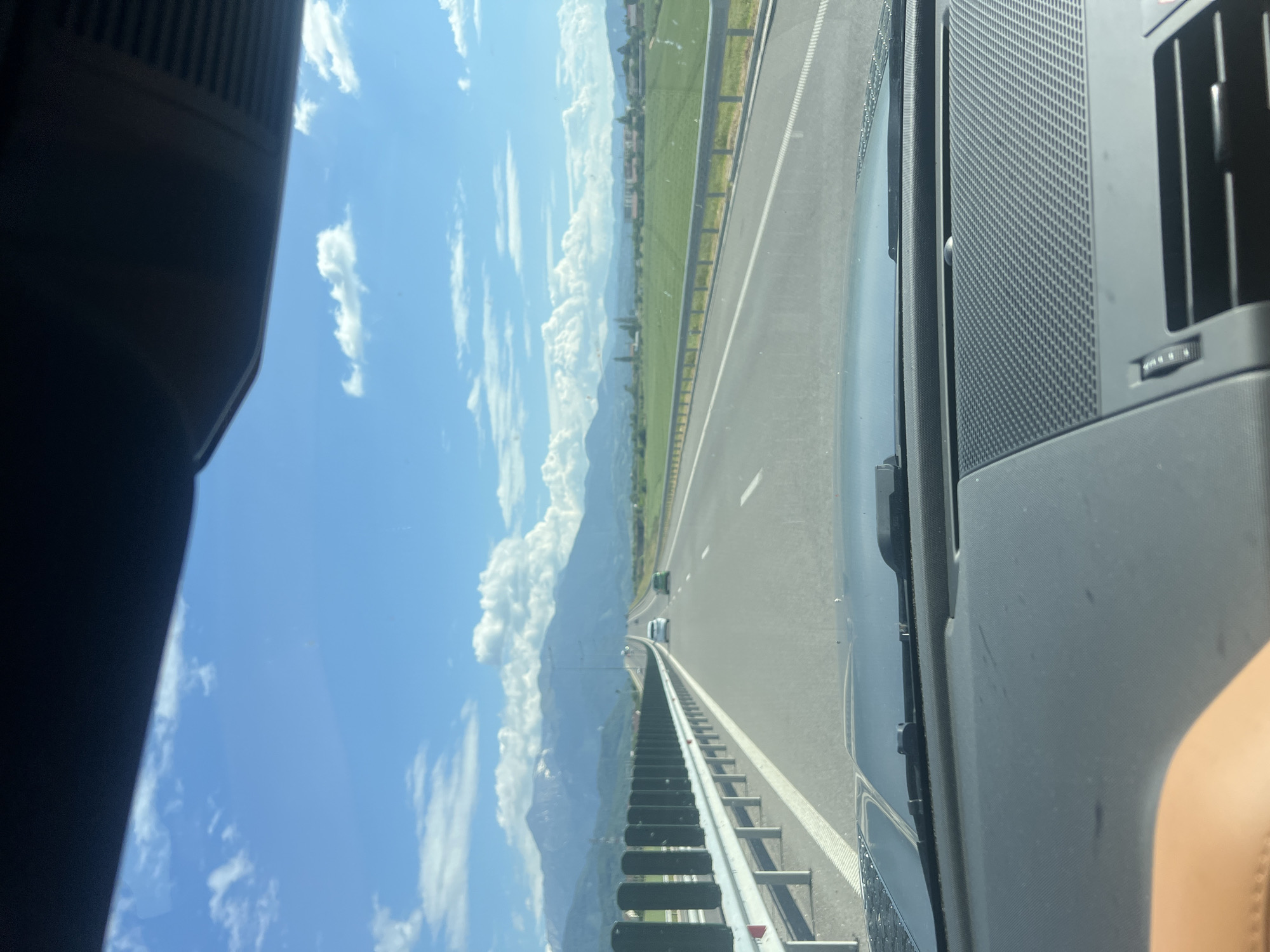 The media file is a photograph taken from the passenger seat of a vehicle, looking forward and to the left. The scene depicts a highway with a clear blue sky and scattered white clouds. In the distance, there are mountains, and to the right of the highway is a green, grassy field. Two cars are visible on the highway. The right side of the photograph shows the interior of the vehicle, including part of the dashboard and an air vent. The sun appears to be shining brightly, suggesting it is daytime. The overall mood is serene and suggests a leisurely drive through a scenic landscape. There is no visible text in the image.