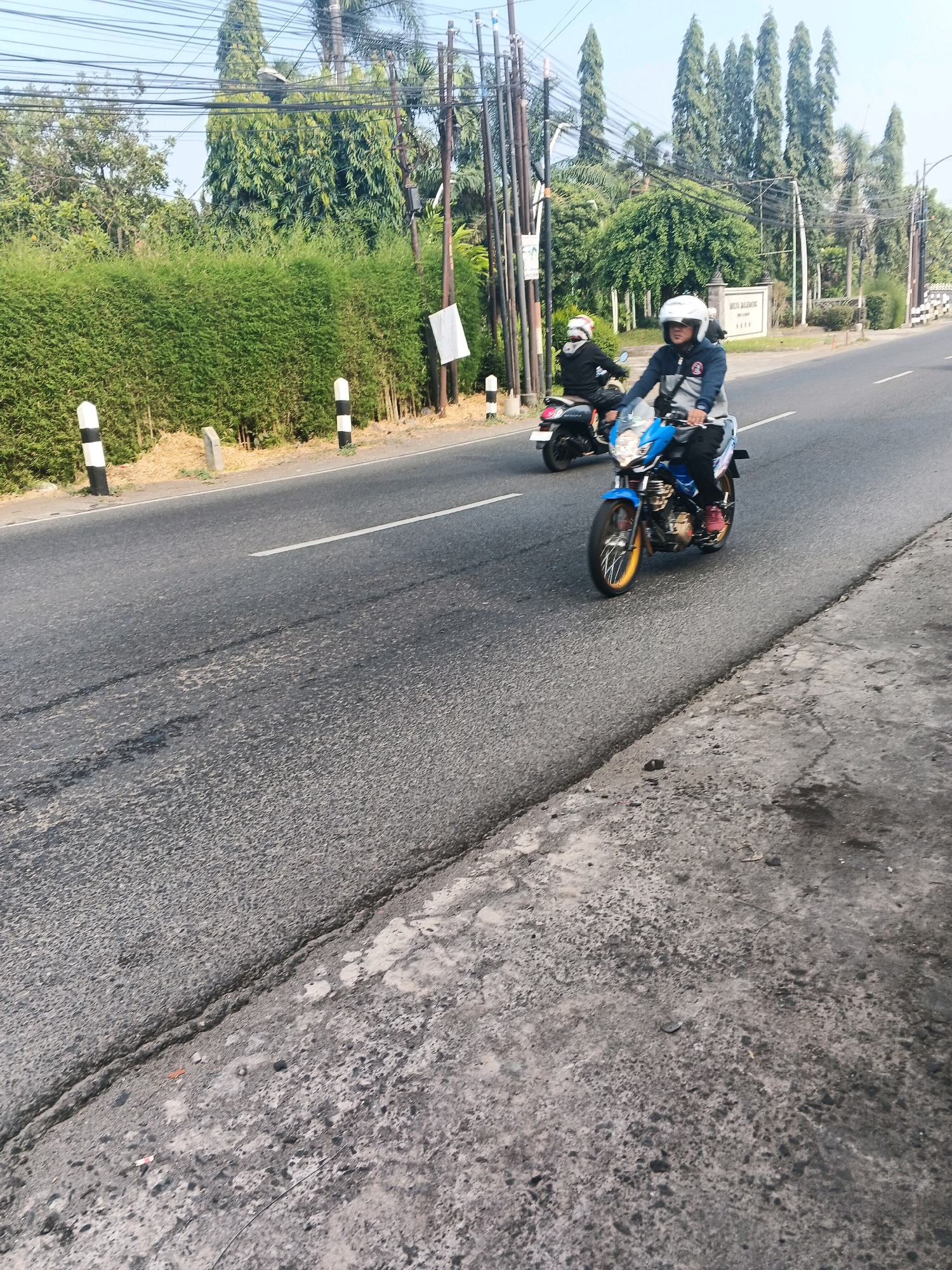 A two-lane asphalt road runs through a suburban area in Ungaran, Indonesia, under a clear blue sky. The road is lined with tall, manicured green hedges on the left and a mix of trees and utility poles on the right. Several black and white striped bollards are positioned along the left edge of the road.
Two individuals on motorcycles are traveling eastbound on the road. The rider in the foreground is on a blue motorcycle with yellow rims, wearing a white helmet, a gray and blue jacket, and dark pants. The rider behind them is on a black motorcycle, wearing a black helmet, a black jacket, and dark pants.
In the background, a white sign with Indonesian text is visible behind the trees. Power lines crisscross the sky, supported by tall utility poles. The road surface shows signs of wear and tear, with some patches of discoloration and a white lane marker. The overall scene depicts a typical day on a road in a tropical urban environment.