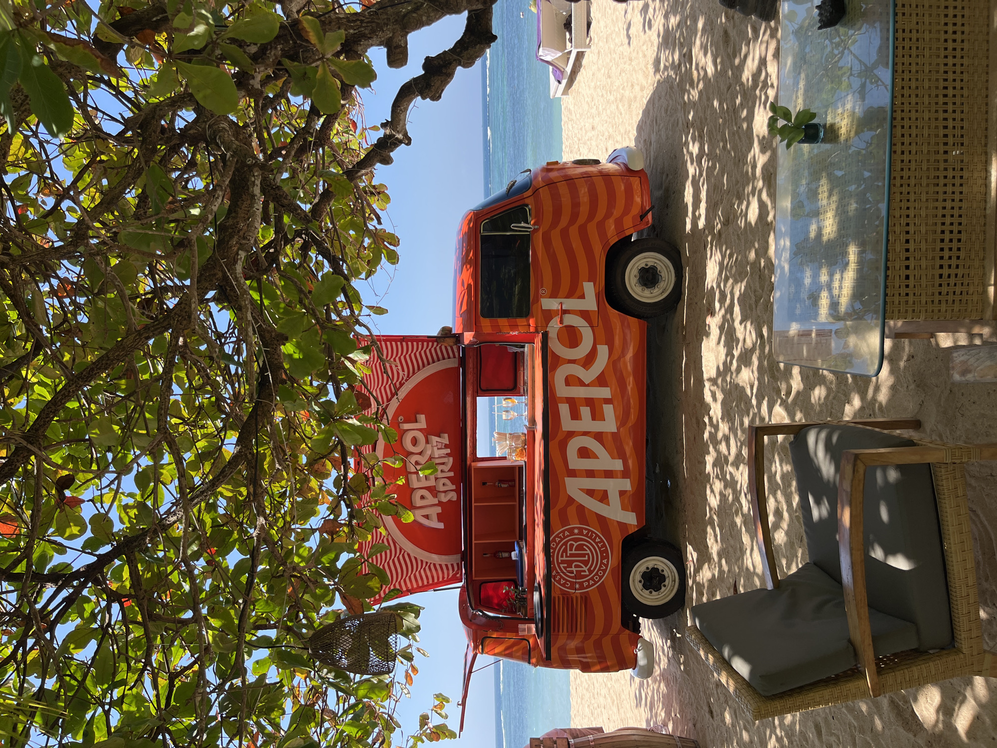An orange and red striped Volkswagen van converted into a mobile bar is parked on a sandy beach in Kuta Selatan, Indonesia. The van is branded with "Aperol Spritz" on a red canopy and the side of the vehicle. The van is open, revealing bottles of alcohol, glasses, and other bar supplies. In the foreground, a wicker chair with gray cushions sits next to a glass-top table with a small plant. The beach is adjacent to the turquoise ocean, with lounge chairs visible in the distance. Lush green foliage from a tree overhangs the scene, casting dappled shadows on the sand. The sky is clear and blue.