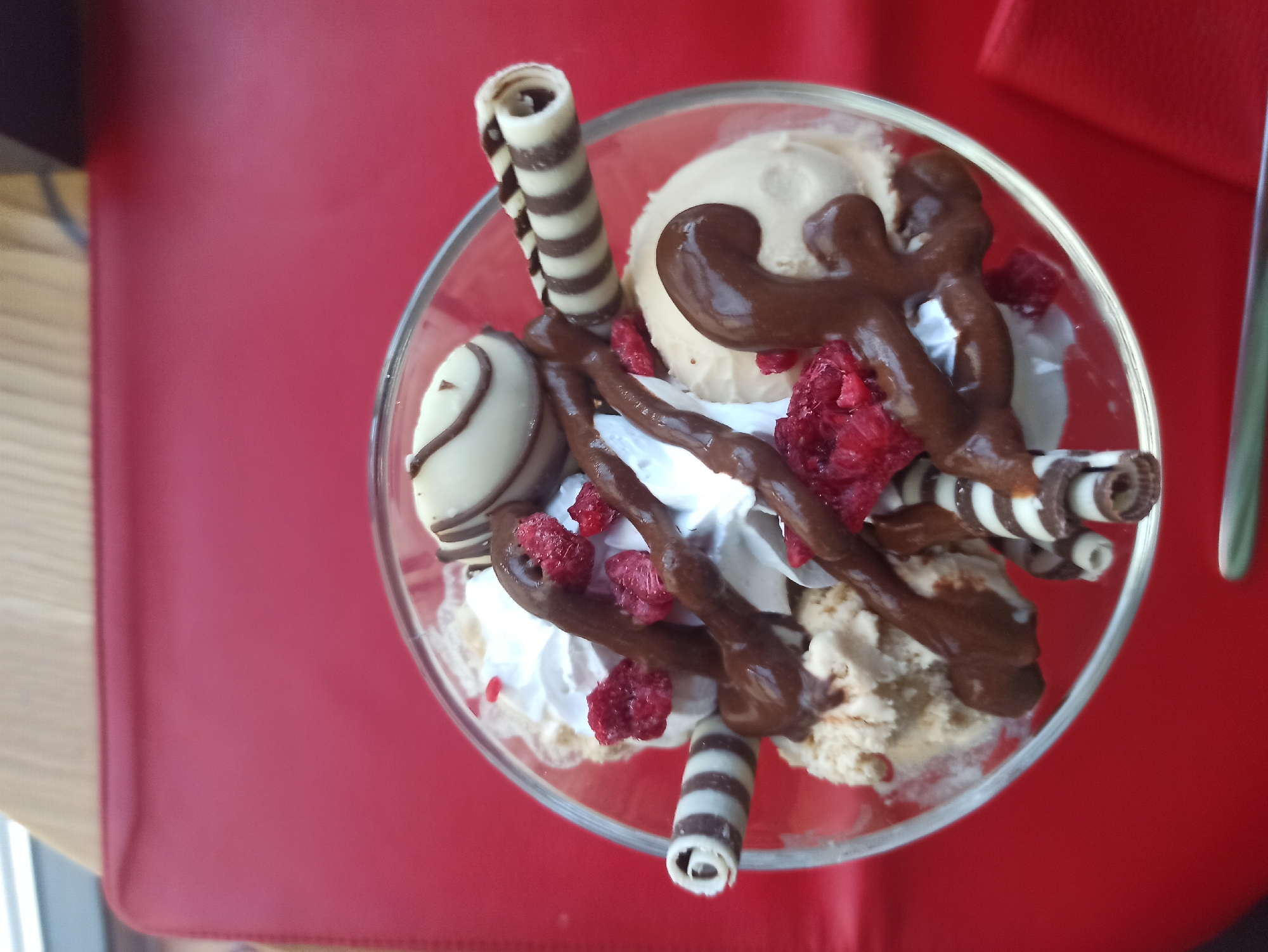 A close-up, overhead shot displays a dessert in a clear glass bowl on a red surface. The dessert appears to consist of ice cream, whipped cream, chocolate sauce, white chocolate balls drizzled with chocolate, striped wafer rolls, and freeze-dried raspberries. The location and specific city are unknown.