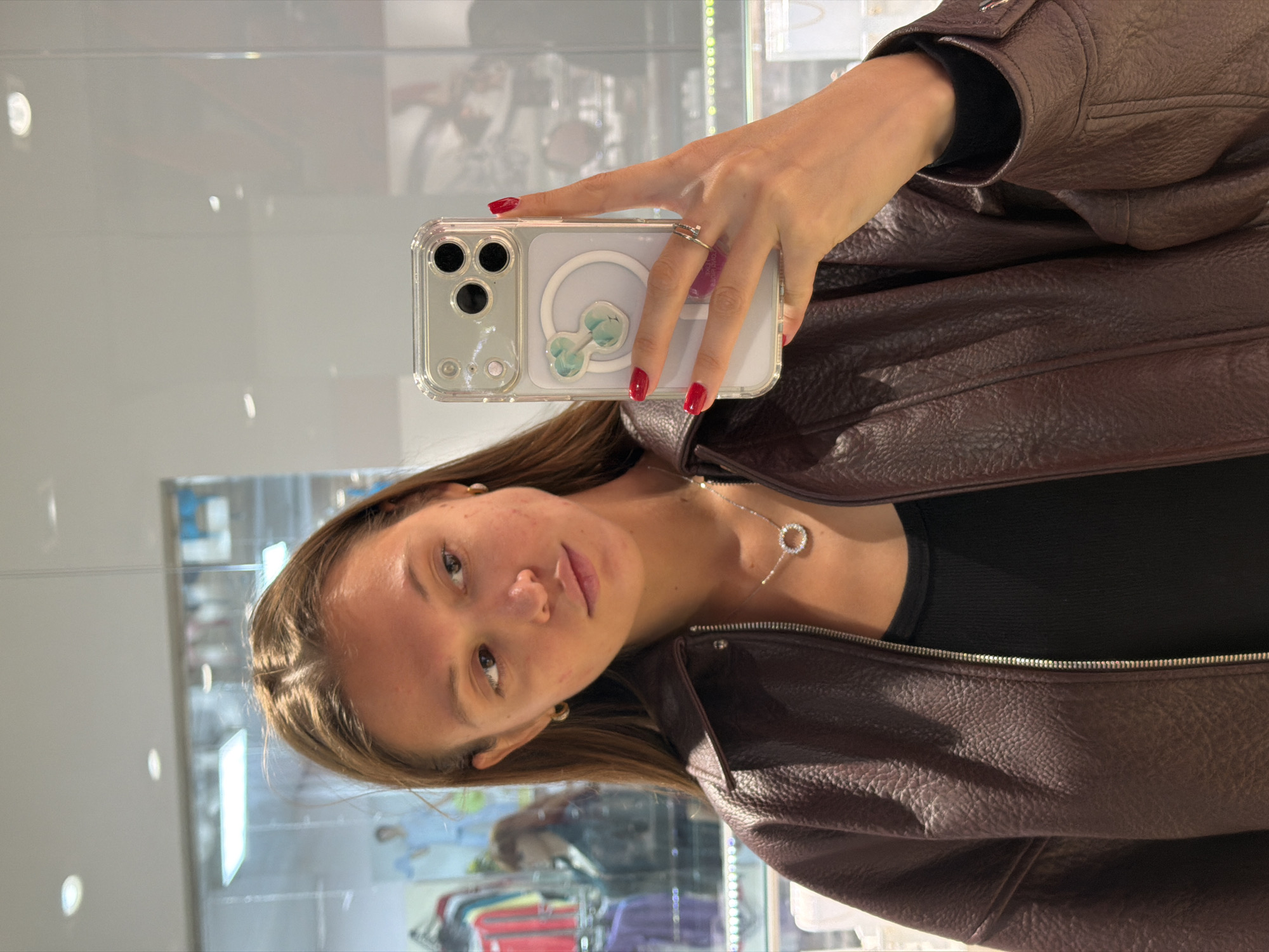 A woman wearing a brown leather jacket is taking a selfie in front of a glass display. She is holding her phone, which has a clear case and a flower sticker on the back. The woman has shoulder-length brown hair, earrings and a necklace. The display behind her appears to be in a store, with clothes and other items visible. It's not possible to confirm the location in the city of Nakhodka, Russia from the image.