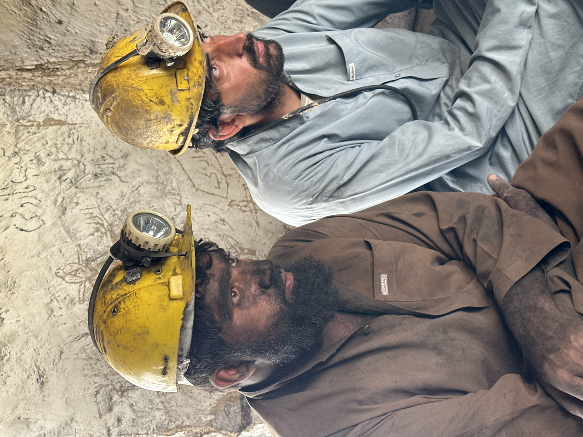 Two men, wearing dusty yellow hard hats with headlamps, sit close together in a dimly lit, rough-hewn environment. The man on the left has a dark, full beard and is covered in dirt, particularly on his face and hands. He wears a brown shirt. The man on the right has a shorter beard and appears cleaner. He wears a light blue shirt. Both men are looking away from the camera, their expressions serious. The background is a textured, dirty wall, possibly inside a mine or cave. The scene suggests a break or resting period for the two men after working in a harsh, dusty environment. This scene is likely in Quetta, Pakistan, a region known for coal mining.