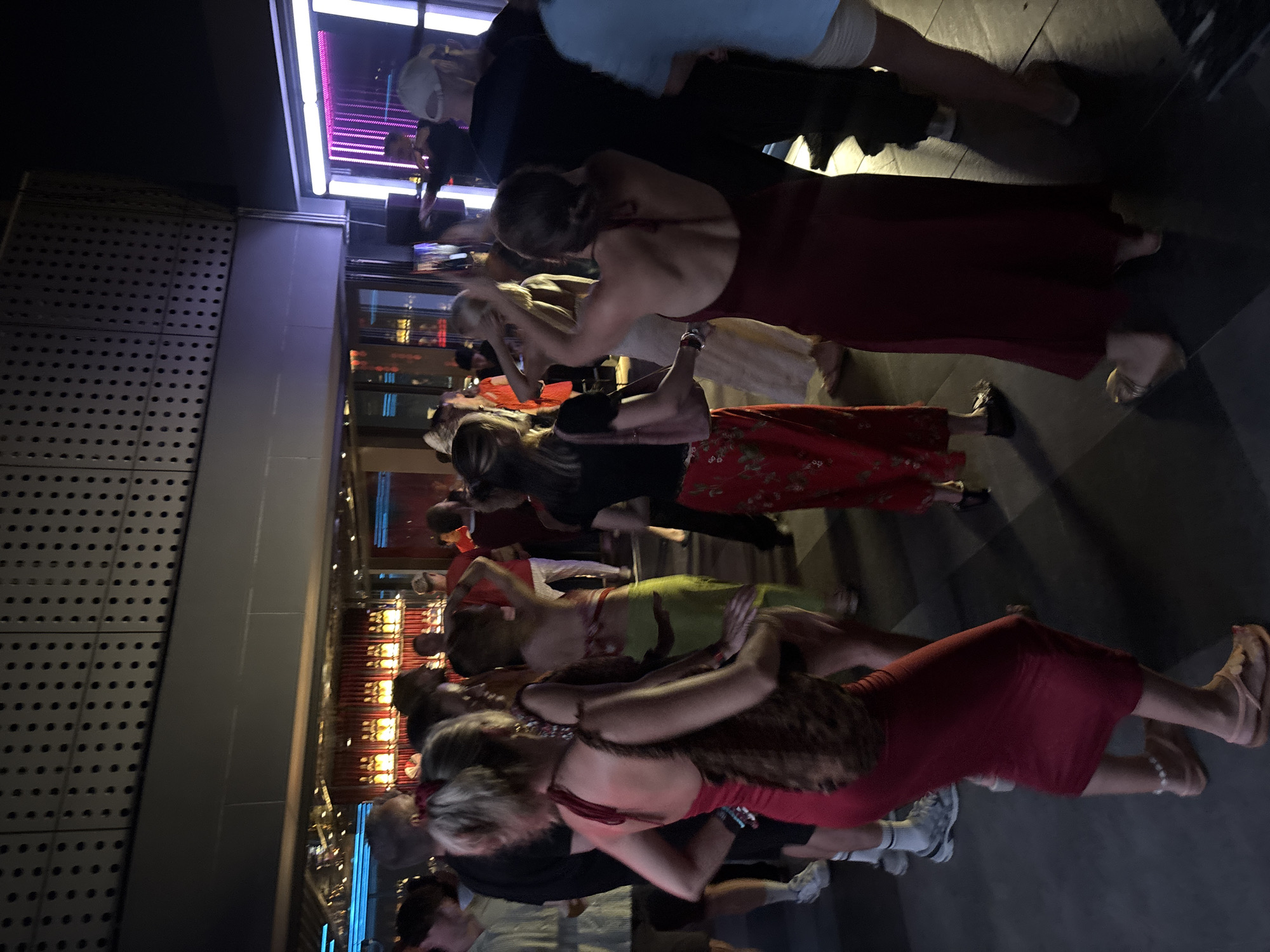 The image depicts a group of people dancing at a nightclub or bar in Đà Nẵng, Vietnam. The scene is dimly lit, with purple and pink lighting accents visible. In the background, there is a bar area with glowing bottles and a stage or performance area with bright vertical lights. The individuals are dressed in casual and semi-formal attire, and some are captured mid-dance. The floor appears to be dark and possibly tiled. The overall atmosphere suggests a lively social gathering.