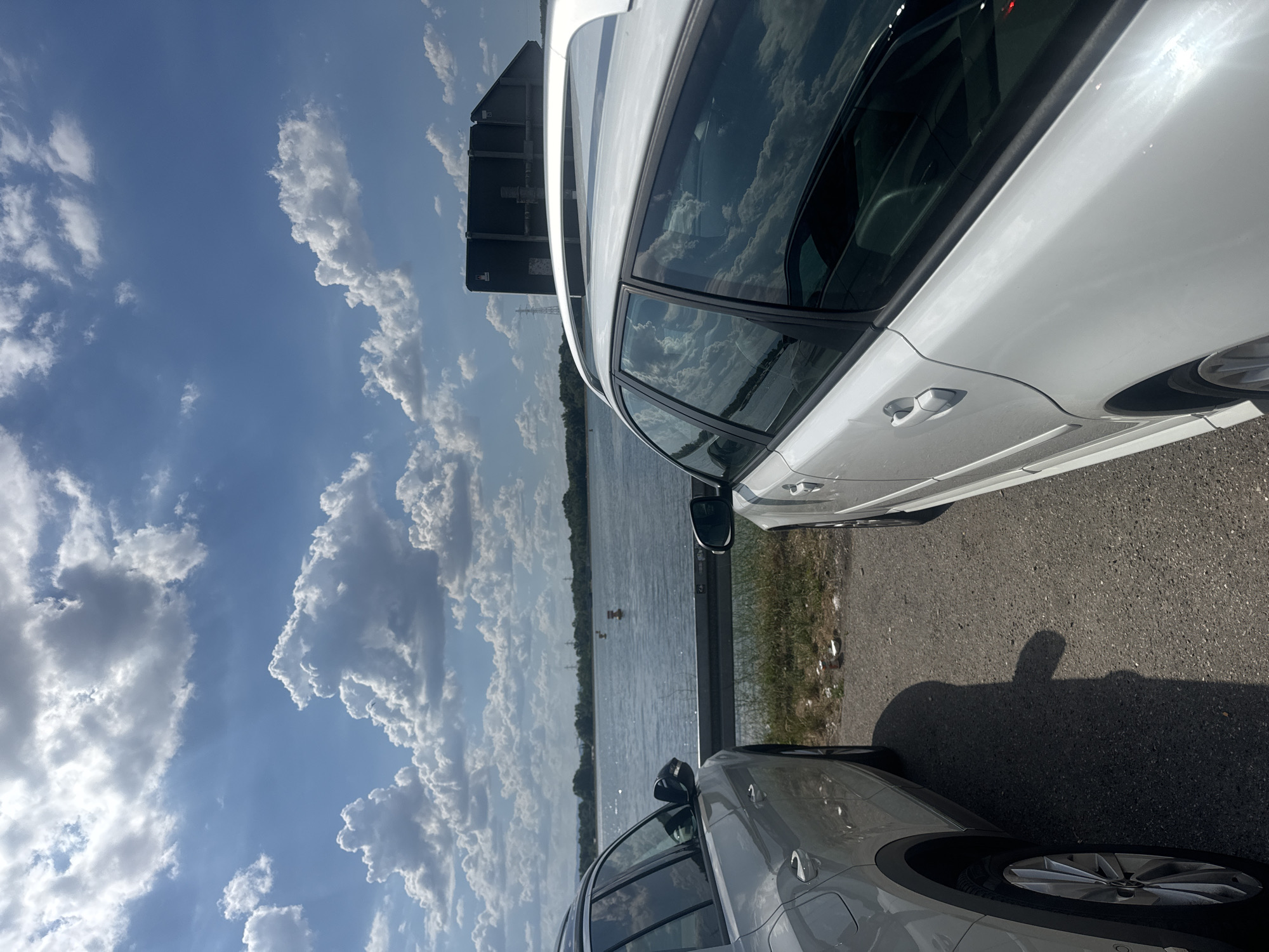 The media file shows a sunlit scene near a body of water, possibly a river or lake, with a backdrop of a bright blue sky filled with dramatic white and grey clouds. Two cars are parked on an asphalt road next to the water. The car in the foreground is a silver SUV, with its front half and wheel visible. The car in the background is a white station wagon, seen from the side, reflecting the sky and clouds in its windows. A shadow of a person, appearing to be making a hand gesture, is cast on the road.
The setting suggests an outdoor recreational area or roadside stop near a natural body of water. The lighting indicates it is daytime, likely mid-morning or afternoon, given the bright sunlight and distinct shadows. The weather appears fair, with abundant clouds but no immediate signs of rain.
The image captures a moment of stillness, with the cars parked and no discernible activity taking place other than the implied presence of the person casting the shadow. The reflections in the car windows add a dynamic element to the scene, mirroring the expansive sky. The overall mood is relaxed and perhaps observational, given the focus on the cars and the surrounding environment. No text is visible in the image.