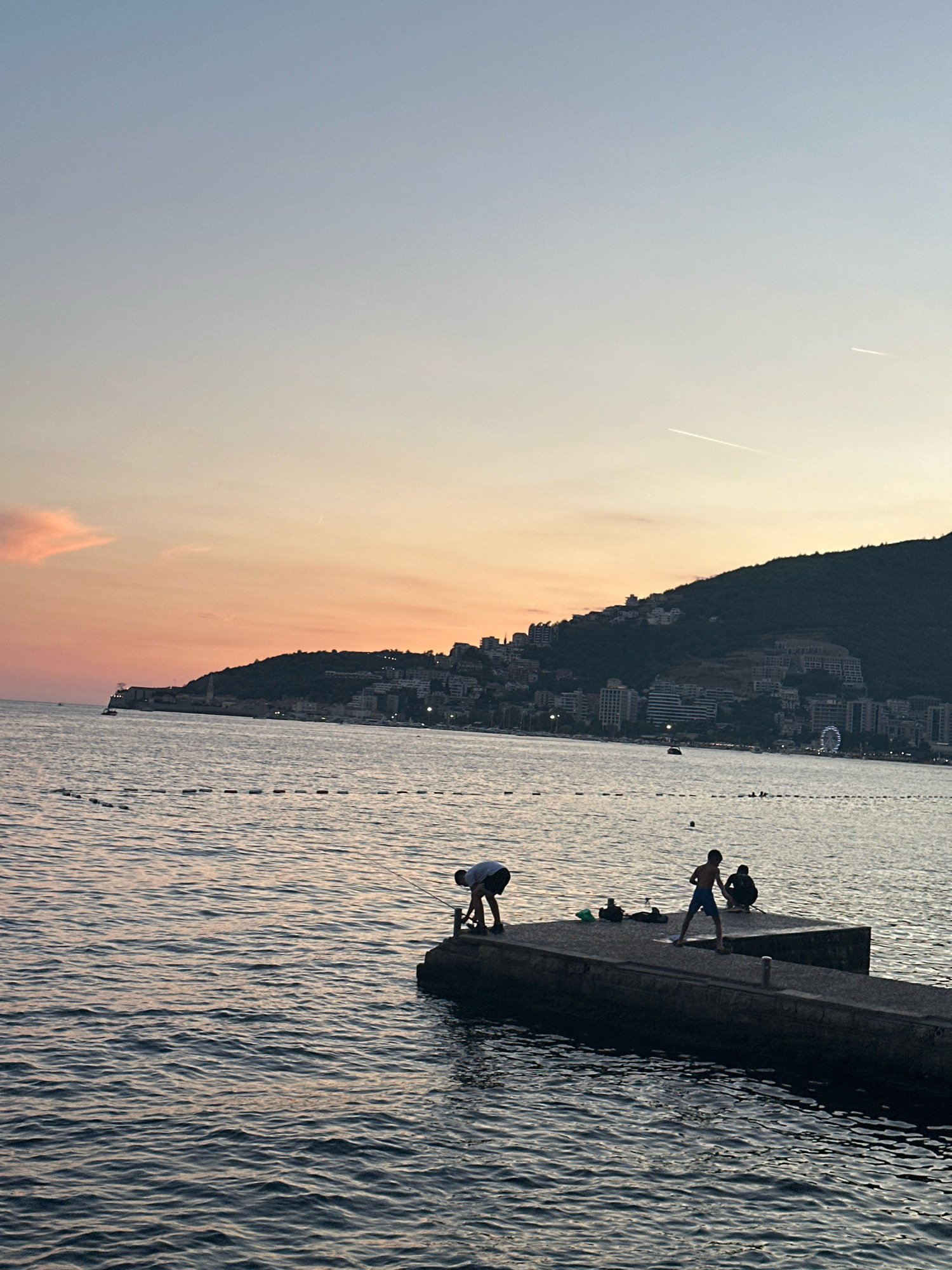 The image captures a sunset over the Adriatic Sea in Budva, Montenegro. The sky is a soft gradient of pale blue to a warm orange, with two faint contrails from airplanes visible. In the distance, a lush green hill is densely populated with buildings, illuminated by the fading light, and a Ferris wheel is visible on the right side of the hill. The sea is calm, with gentle ripples reflecting the colors of the sky.
In the foreground, a concrete pier extends into the water. On the pier, three figures are present. On the left, a person wearing a white shirt and dark shorts is bending over, possibly fishing. In the center, a young person in blue shorts stands with their arms extended, and to the right, another person is seated. Various items, possibly fishing gear or bags, are scattered on the pier near the standing figure. The scene conveys a peaceful evening atmosphere.