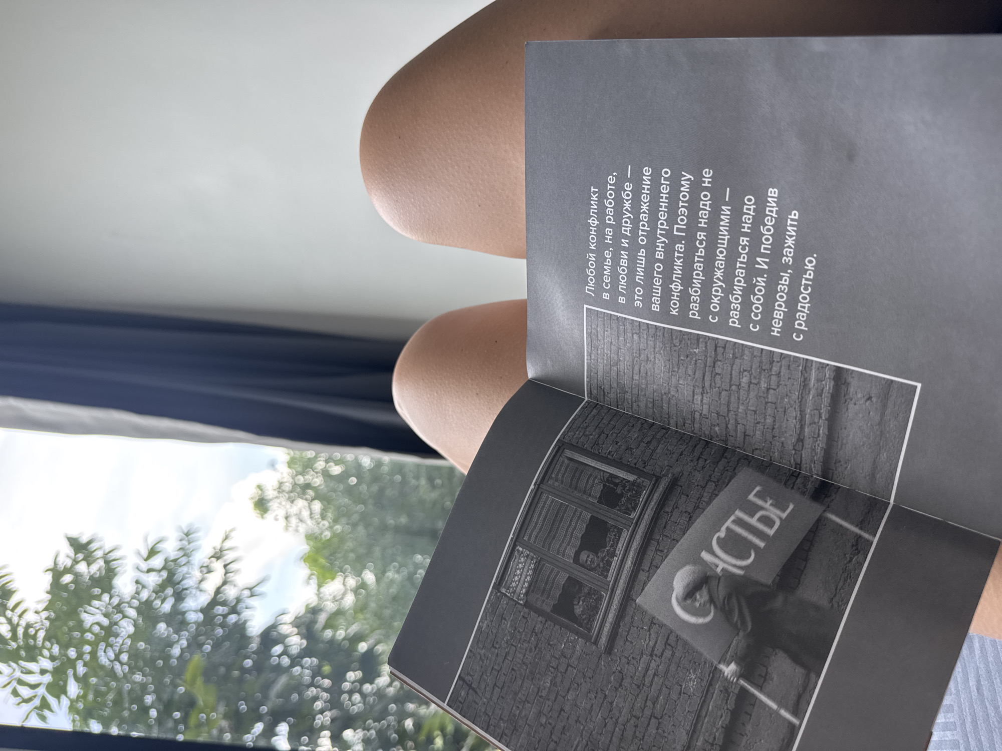 An open book is held by a person whose legs are visible in the foreground. The book contains black and white photographs and text. One photograph shows a person holding a sign with Russian text that appears to say "СЧАСТЬЕ" (Happiness). The text in the book is also in Russian. The background shows a window with trees and greenery visible outside, suggesting an outdoor or natural setting. The image does not provide any information about the location being in the city of Ratsada, Thailand.