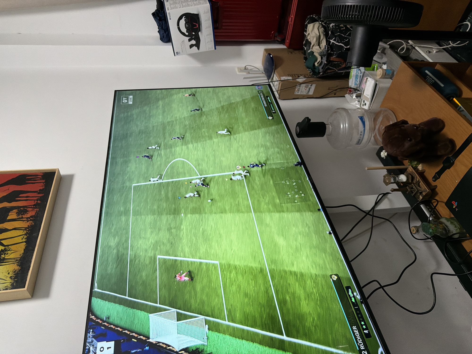 This media file captures an indoor scene, likely a room in a residence within Jiangning District, China. A large flat-screen television, rotated 90 degrees clockwise, dominates the left side of the frame, displaying an ongoing soccer (football) video game. The game screen shows a green pitch with white markings, a goal, and numerous player avatars dressed in white and darker kits, with a goalkeeper in pink. Visible on-screen text from the game interface includes "LIVE," the score "0-0," player names such as "RÜDIGER 2" and "MBAPPÉ 7," along with other small icons and numbers.
To the right of the television, a wooden desk or shelf is cluttered with various objects. These include a black desk fan, a clear plastic water jug with a black cap, a brown stuffed animal, and a blue JBL speaker. Several cardboard boxes are stacked, with one prominently displaying "Thrustmaster" branding, suggesting a gaming steering wheel. A PlayStation console with its logo is also visible. Loose black cables and wires are strewn across the surface. On the white wall to the left of the TV, a framed picture depicting a reddish-orange and black scene, possibly giraffes, is hanging. The room appears to be illuminated by artificial light.