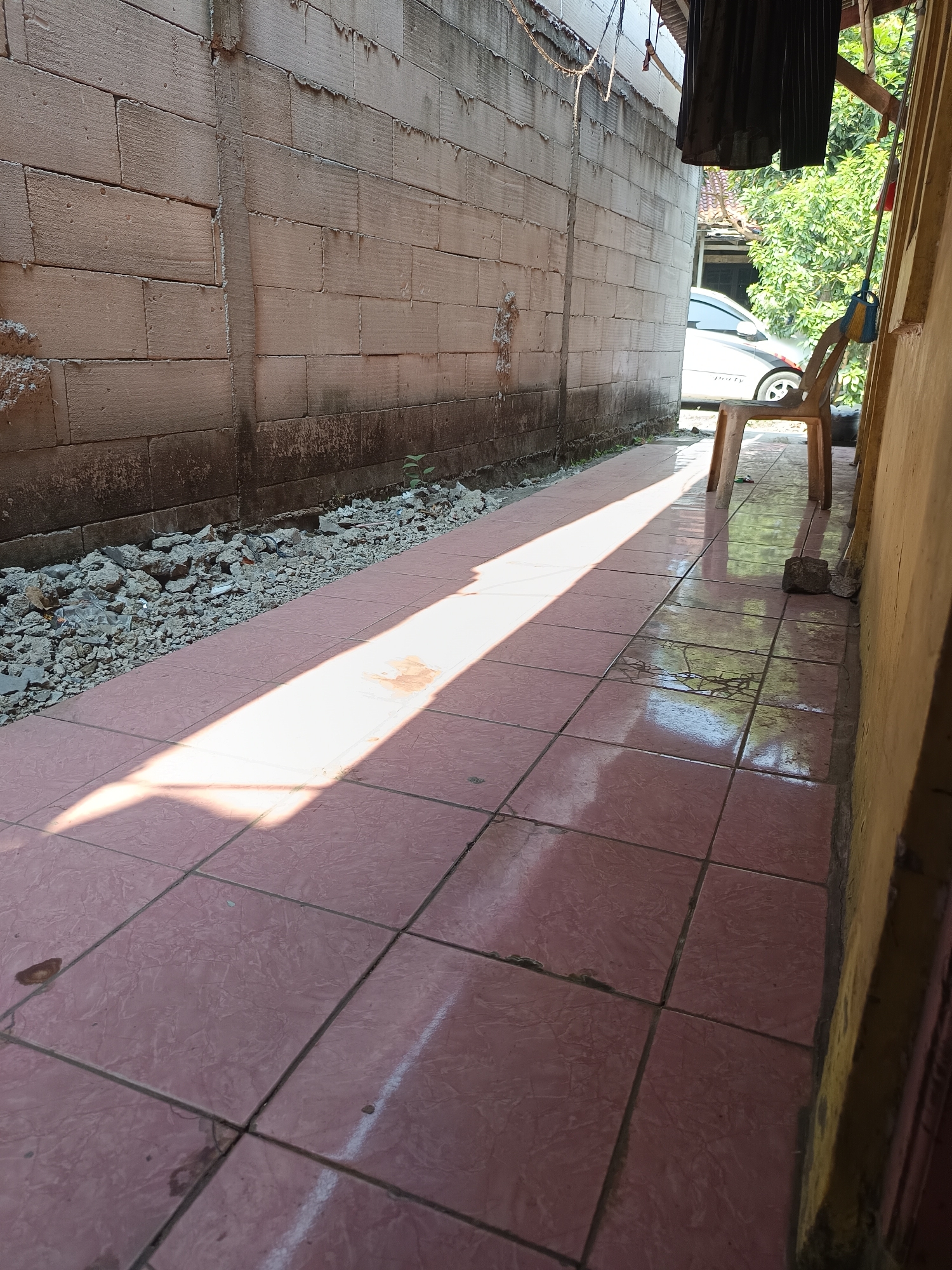 A narrow outdoor walkway is depicted, bordered by a rough, light-colored brick wall on the left and a yellow-painted structure on the right. The walkway is paved with pinkish-brown square tiles, some of which appear cracked or stained. Sunlight streams across the tiles, creating a bright diagonal band of light. To the left of the walkway, there is a strip of gravel and debris along the base of the brick wall. In the background, a silver car is visible, parked next to a light brown plastic chair. A broom with a blue and yellow brush head leans against the chair. Lush green foliage is present behind the car. A dark piece of fabric hangs from an overhead structure. The scene appears to be an alleyway or side passage within a residential area in Talaga Sari, Indonesia.