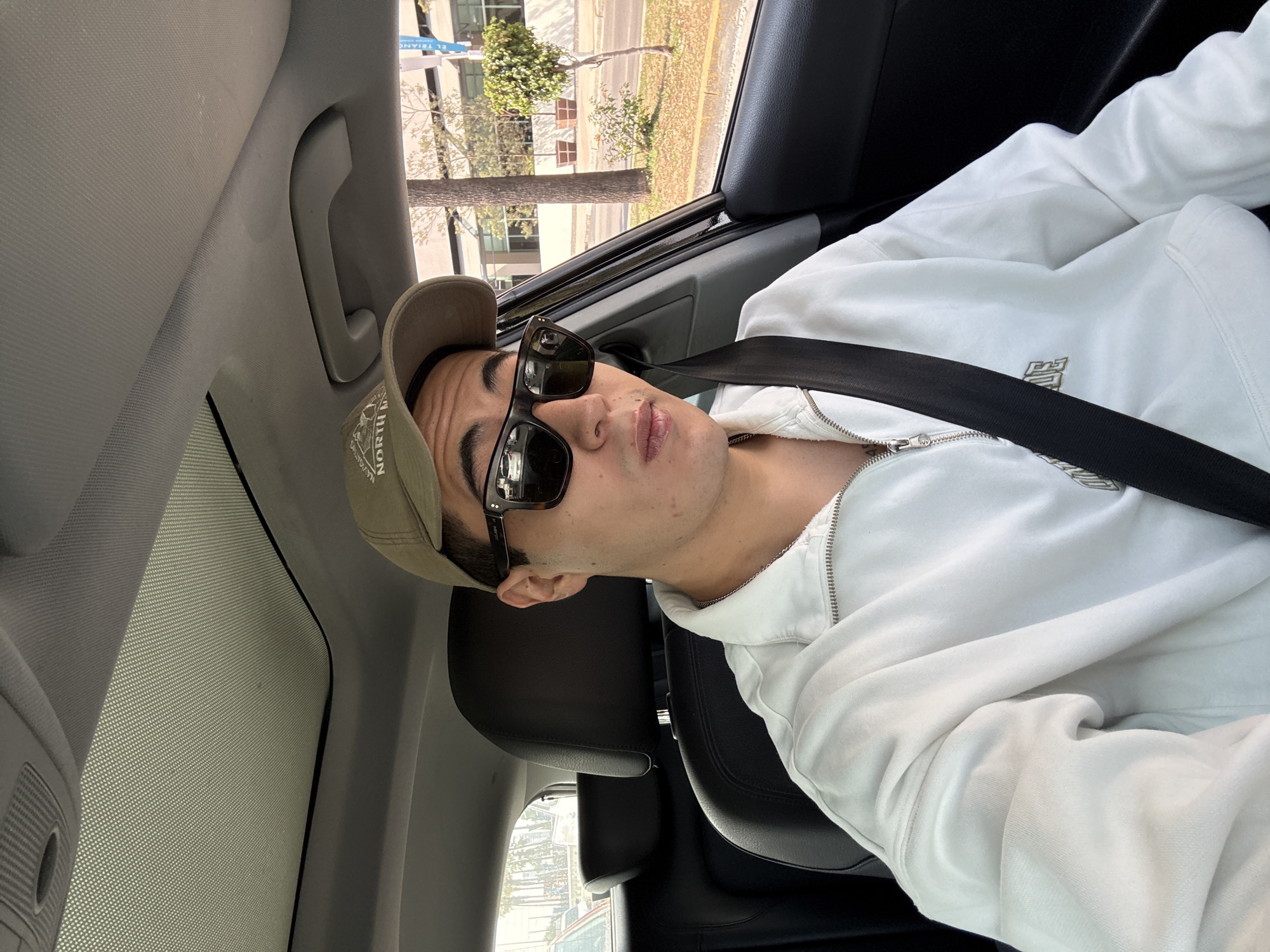 A young man is the central subject of this image, captured in a selfie-style shot from a slightly low angle, taken inside a car. He is seated in the front passenger seat or possibly the driver's seat, looking towards the camera with a neutral, relaxed expression. He wears dark, rectangular sunglasses and a beige or khaki baseball cap with some faint, dark text or a logo on the front that appears to say "NORTH B...". His attire includes a white zip-up hoodie, which has a subtle, light-colored embroidered logo or emblem on the left chest area. A black seatbelt crosses diagonally over his chest.
The setting is the interior of a car, with parts of the headliner, a grab handle, and the seat visible. Through the car windows, a sunny daytime scene is visible, suggesting good weather. Outside, there are trees, buildings, and a road or pavement. A notable detail outside the car is a blue sign, partially visible, which clearly shows the text "EL TRIANGULO". Given the location context of Puebla, Mexico, "El Triángulo" likely refers to a local landmark or shopping center. The car appears to be stationary or moving slowly, allowing for a clear view of the outside environment. The sunroof, or moonroof, is also visible, with a mesh-like patterned shade covering it. The overall mood is casual and relaxed.