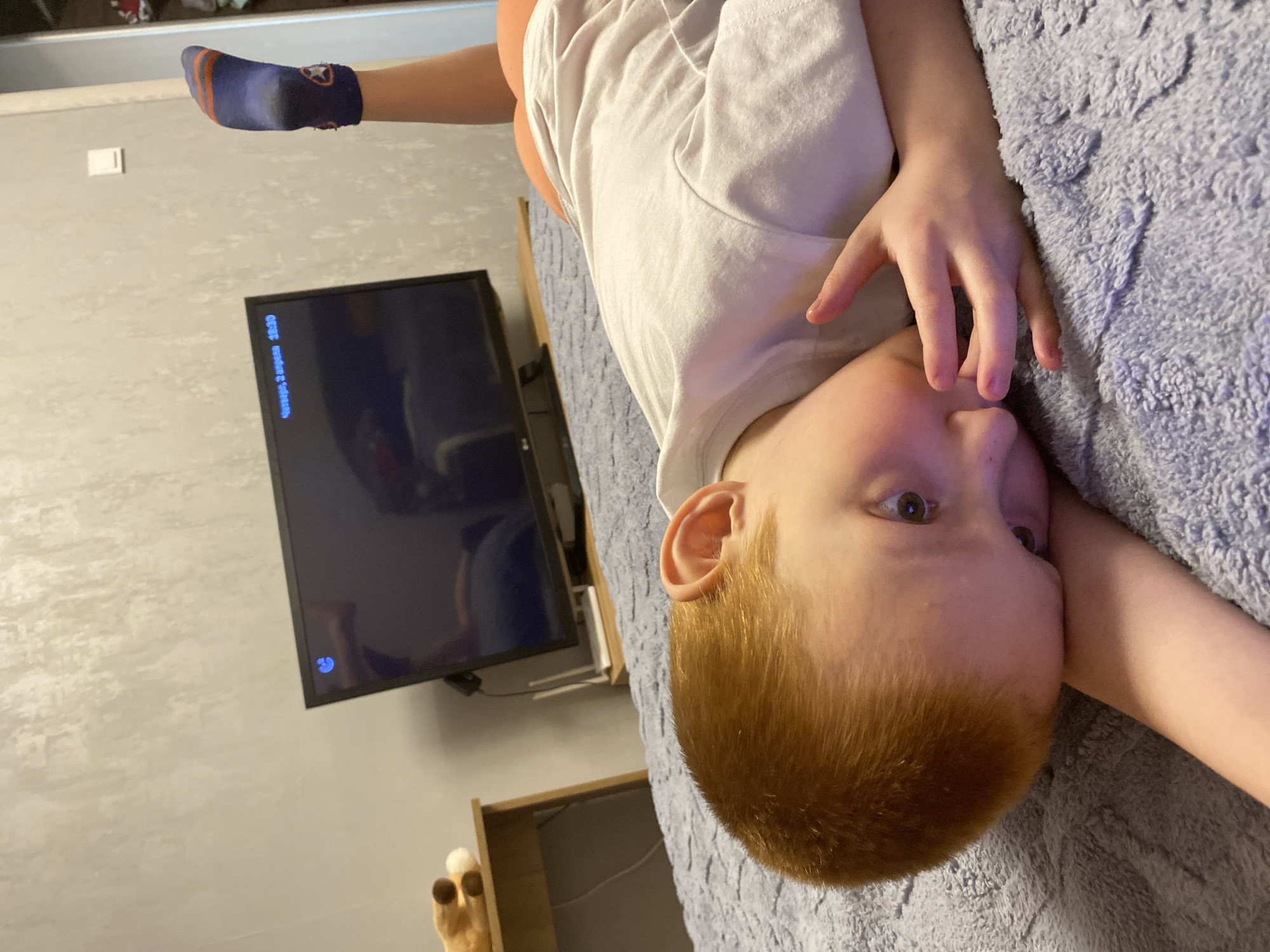 A young child with red hair lies on a gray, textured blanket. The child is wearing a white t-shirt. The child's hand is near their face. The child is wearing a sock on their foot. A television is on the wall. The city of Noviy, Russia is not discernible from the image.
