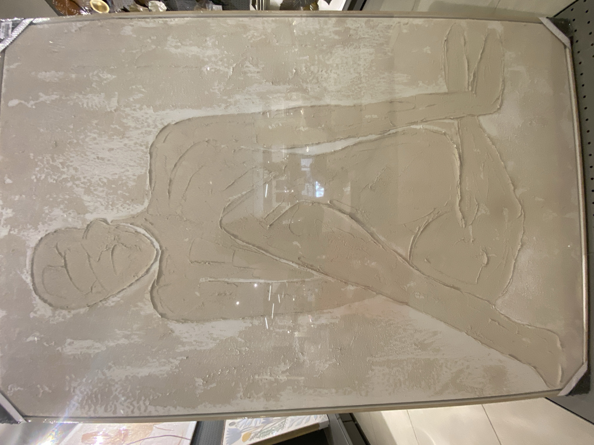 This is a media file of a framed textured artwork. The artwork is a bas-relief sculpture depicting a reclining figure. The figure is rendered in a neutral, earth-toned material with a rough, textured surface. The body is elongated and simplified, with distinct lines suggesting limbs and the torso. The head is a rounded mass with subtle indentations for features. The background of the artwork is also textured, with lighter, more irregular patches of material that create a sense of depth and contrast. The artwork is framed with a thin, metallic-colored border, and the corners of the frame are reinforced with small, dark triangular pieces. The overall aesthetic is abstract and minimalist, focusing on form and texture. The lighting in the file reflects off the glossy surface of the frame, creating bright highlights and obscuring some of the finer details of the artwork itself. The scene appears to be an indoor setting, possibly a gallery or home, given the framing and the hint of other objects in the background. There is no visible text in the media file.