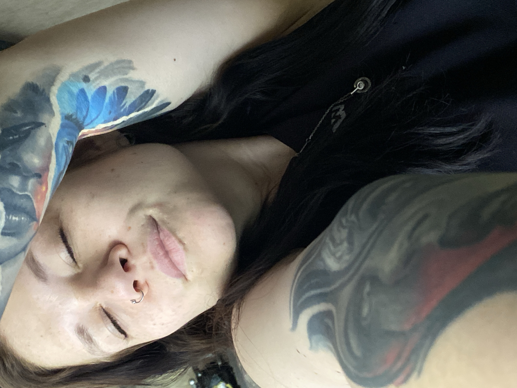 A young woman with tattoos on both arms lies on her back. She has dark hair, closed eyes, and a nose ring. She is wearing a black top with a chain. The image does not provide enough information to determine if she is in the city of Chaikovsky, Russia.