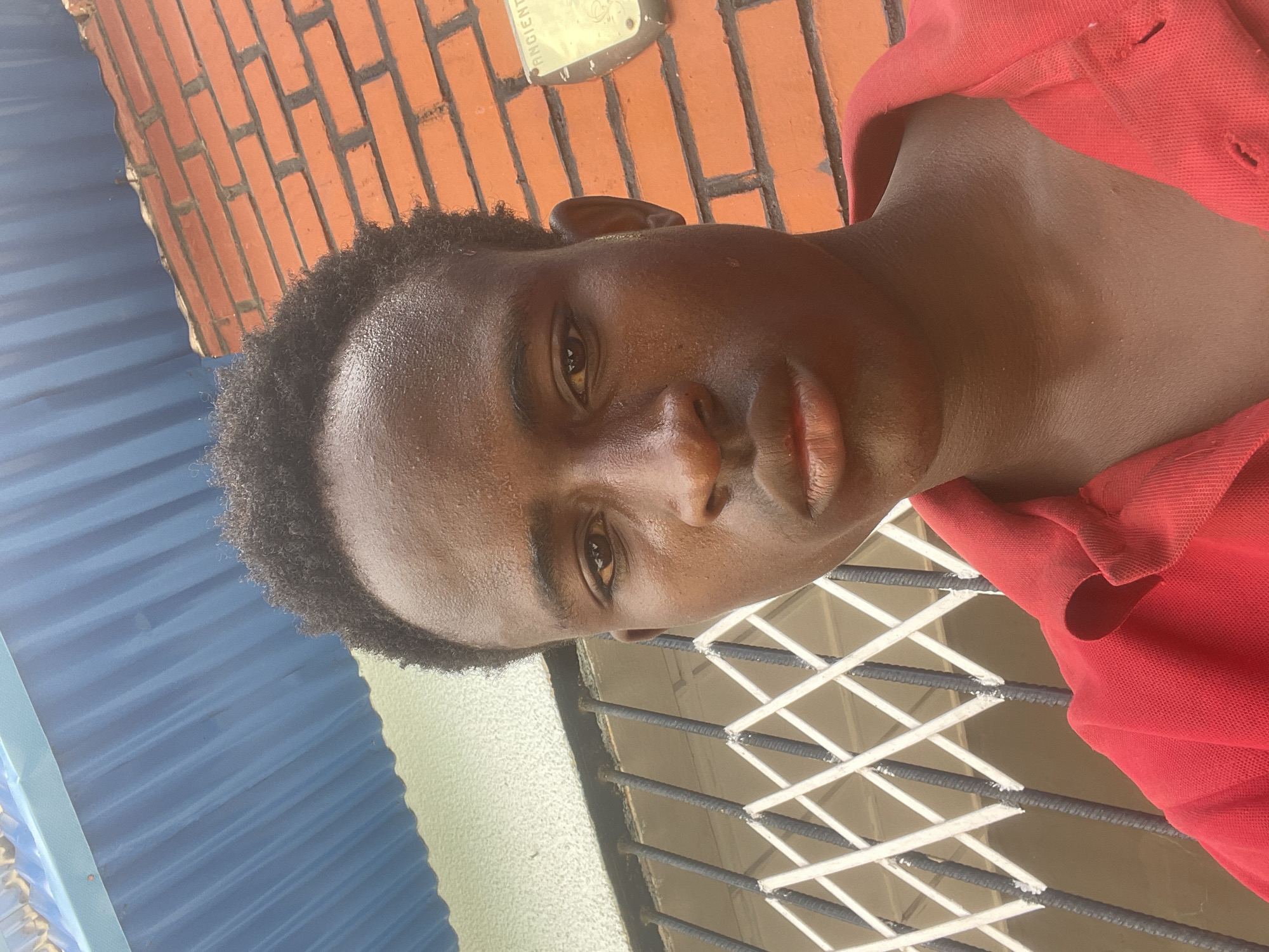 The image is a close-up selfie of a young Black male in Jos, Nigeria, with a red collared shirt. The background features a blue corrugated metal roof, a white and black metal gate, and an orange brick wall with a light-colored fixture labeled "ANCIENT." The lighting suggests it is daytime, and the subject appears to be outdoors.
