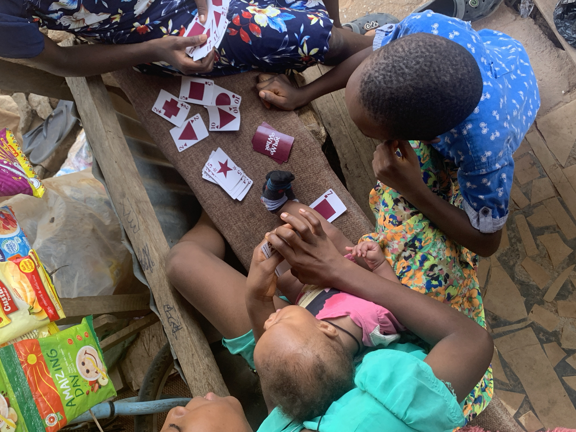 Here is a detailed description of the image:
**Scene:** The image depicts a group of people playing cards on a makeshift table outdoors. Based on the packaging of food products present, it is likely that the location is in Nigeria, possibly in the city of Sabon Tasha.
**People:** Several individuals are visible. One person is holding a baby in their arms, while others are seated around the table, engaged in the card game. The subjects' clothes are colorful and patterned.
**Subjects:** The focus of the image is the card game. Playing cards are scattered on the surface of the table.
**Objects:** Various objects are visible, including the playing cards ("What Next" brand visible), snack packaging, a baby sock, clothing, and a makeshift wooden table.
**Actions:** The primary action is playing cards. Individuals are holding cards, and some are laid out on the table. The interactions among the people appear casual and friendly.
**Environment:** The surroundings appear to be somewhat cluttered and possibly outdoors. There is a mix of textures and colors, suggesting a busy and dynamic environment.
The location appears to be in a low-income area.
