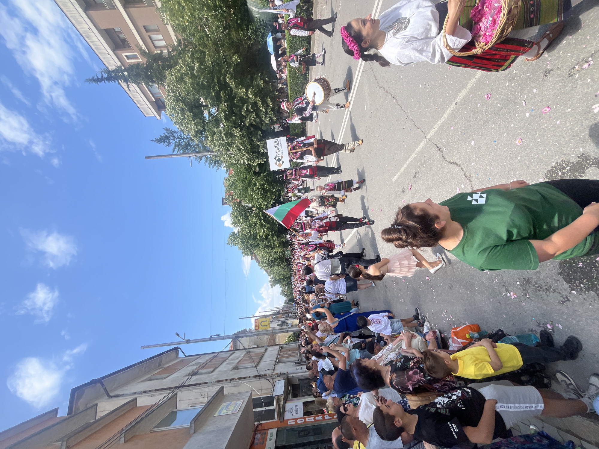 This image captures an outdoor daytime parade or festival unfolding on an asphalt street in an urban environment. The street is lined with spectators on the left, observing a procession of individuals, many dressed in traditional Bulgarian folk costumes.
In the immediate foreground, to the right, a woman in a white blouse, traditional skirt, and a pink floral headpiece holds a wicker basket filled with pink rose petals, some of which are scattered on the street. To her left, a woman in a green t-shirt looks towards the parade.
The parade features numerous participants marching away from the viewer. A Bulgarian flag is prominently displayed on the left side of the procession. Further along, a banner is held aloft, reading "ТРАКИЙЦИ" (Thracians) and "Клуб за народни танци" (Folk Dance Club) with a geometric logo. Several participants wear distinct red or black embroidered vests over white shirts, dark trousers, and traditional hats. One male participant is actively playing a large, double-headed drum (tupan) with two sticks. Children in miniature versions of the traditional costumes are also part of the parade.
On the left side of the street, a diverse crowd of spectators, ranging from children to adults, stands along the curb, many raising their hands or holding up phones to capture the event. Behind them are multi-story buildings, some appearing older with exposed concrete and varying window designs, and others more modern. Commercial signs, including "BILLA" and "Подаръци" (Gifts), are visible on these buildings.
On the right side of the street, lush green trees with dense canopies line the thoroughfare, partially obscuring more distant buildings. The sky above is clear and bright blue, dotted with scattered white cumulus clouds, indicating sunny weather. The event appears festive and well-attended. The location is an unidentified street in an unknown city.