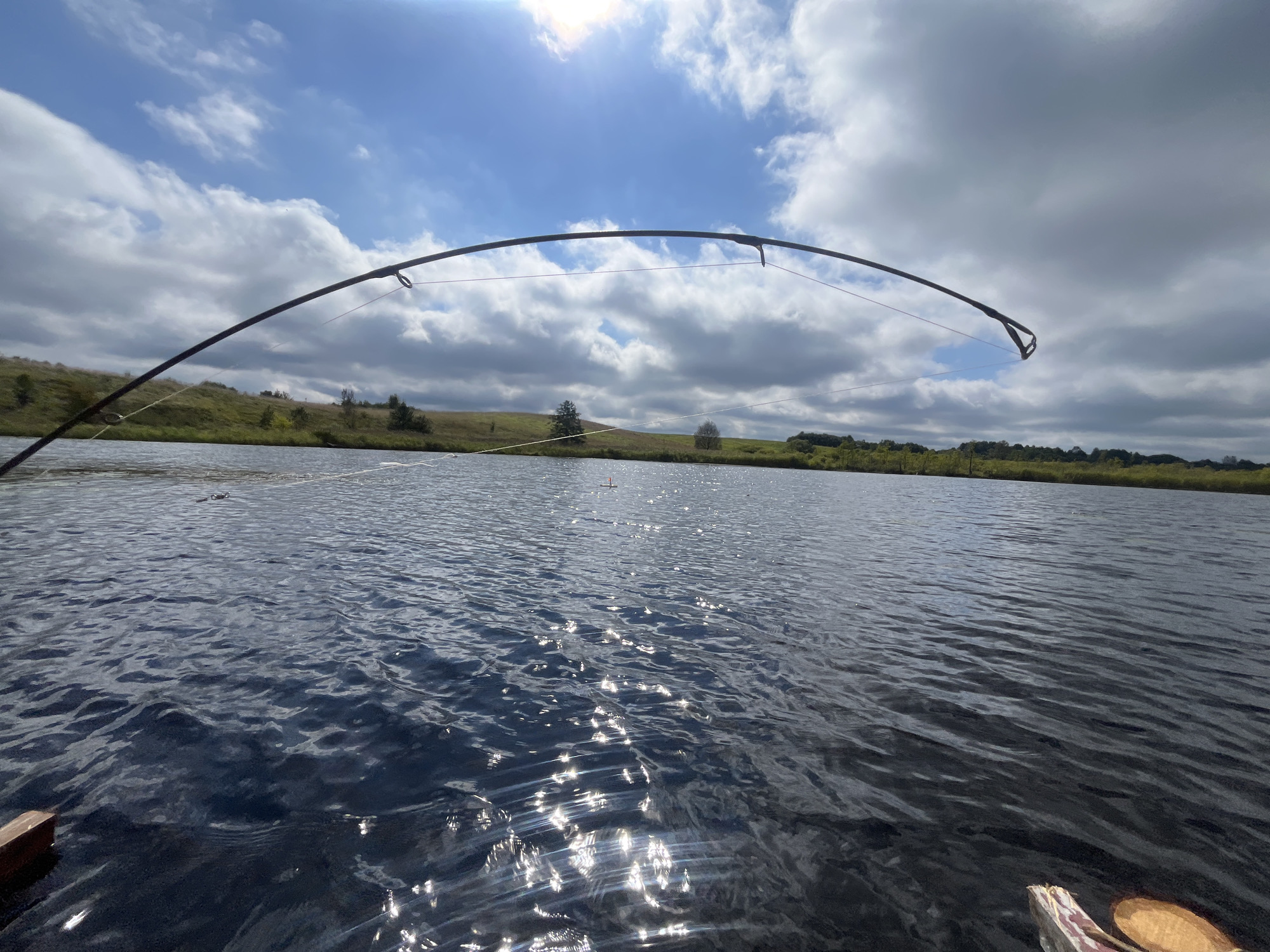 An angled, low-angle shot of a serene lake on a cloudy day, with a fishing rod arching from the foreground into the frame. The water is a deep blue, reflecting the sky and sunlight in shimmering patterns. The shoreline, visible in the background, is covered with green grass and scattered trees, with a gently sloping hill. The sky is a mix of bright blue patches and large, fluffy white clouds, with the sun peeking through, creating lens flares. A few small, orange buoys are visible in the distance on the water. The image appears to be taken from a boat, with a portion of a wooden oar visible in the lower right corner. The scene is peaceful and natural, evoking a sense of calm and outdoor recreation. The location is not specified in the image or the provided context.