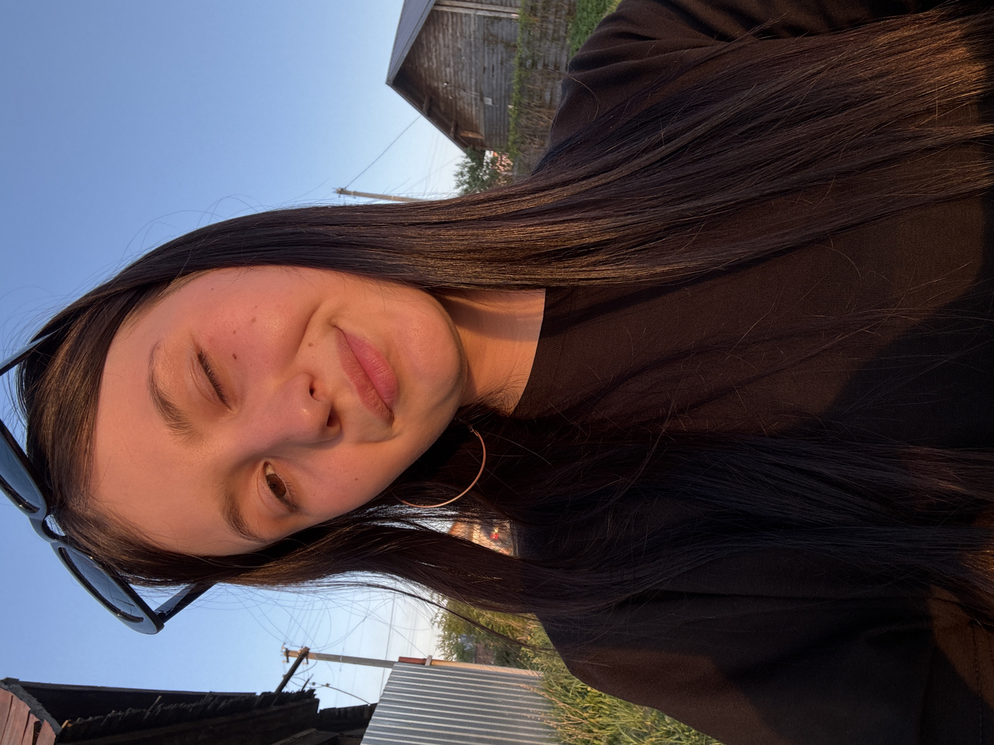 A young woman with long, dark hair is winking in a selfie outdoors. She wears large hoop earrings, a dark shirt, and sunglasses are perched on her head. The background includes an old wooden building and glimpses of a zinc or corrugated metal structure. The sky is a clear, light blue. The scene appears to be illuminated by the setting sun, with a warm glow on the woman's face. The location may be Фоки, Russia.