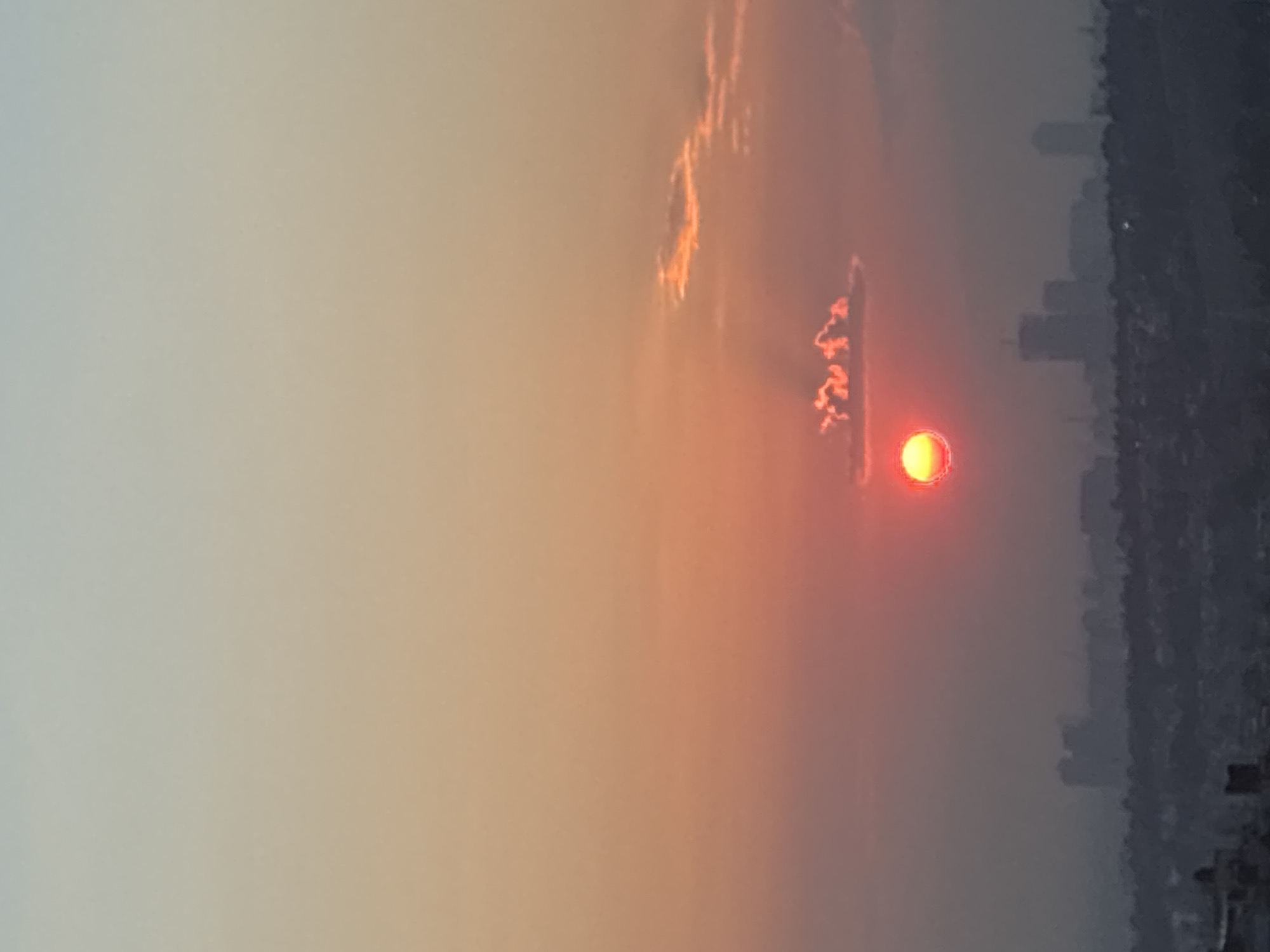 The image captures a sunset over Huixquilucan de Degollado, Mexico. The sun appears as a red-orange disk, partially obscured by haze and clouds. The sky exhibits a gradient, transitioning from a lighter shade near the horizon to a darker, more muted tone higher up. The silhouette of a city skyline can be discerned in the distance. The atmosphere appears smoky or dusty, contributing to the hazy appearance.