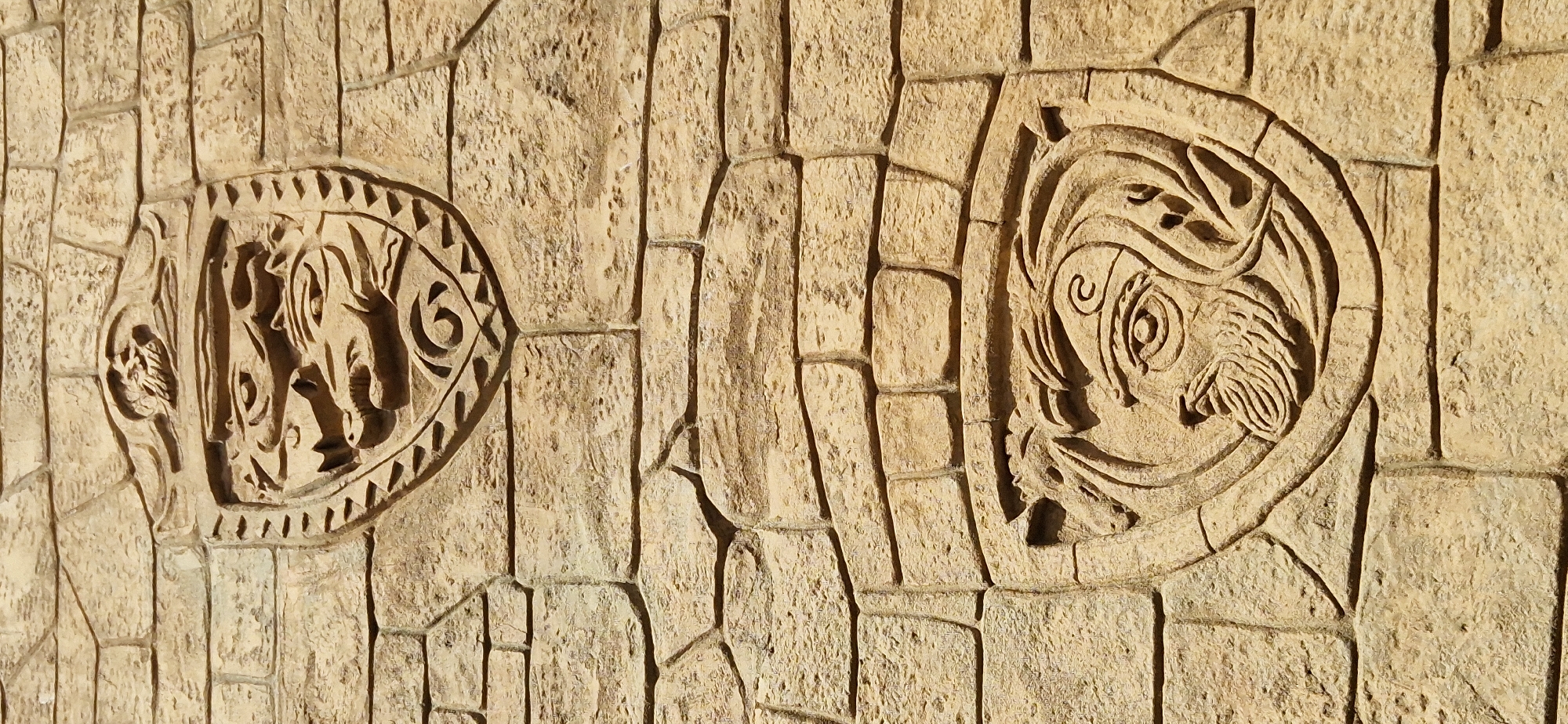 The image displays a section of a stone wall adorned with two carved circular reliefs. The wall itself is constructed from irregularly shaped, light-brown stones, meticulously fitted together to form a textured surface. The carvings are centrally located on two of these stones, appearing as intricate, shallow reliefs.
The left relief depicts what appears to be a stylized animal, possibly an elephant, with large ears and tusks. Its form is outlined by a decorative border featuring triangular motifs and a circular swirl. The right relief features a face with large, prominent eyes and flowing, stylized hair or headdress. This relief also has a circular border, albeit with a simpler, more abstract design. Both carvings have a similar color to the surrounding stone, suggesting they are made from the same material and have weathered over time.
The lighting in the image suggests it is daytime, with a warm light casting subtle shadows that highlight the texture of the stone and the depth of the carvings. There are no visible people, text, or specific cues to indicate the weather or a precise geographical location. The scene is static, focusing solely on the architectural detail of the wall and its ornamental elements. The overall impression is one of ancient artistry and enduring craftsmanship.